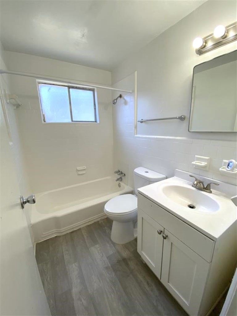 A clean, bright bathroom featuring a bathtub with shower, a white toilet, and a sink with a cabinet underneath. There is a large mirror above the sink and natural light coming through a window. The floor is tiled in a light wood pattern, contributing to a modern aesthetic.