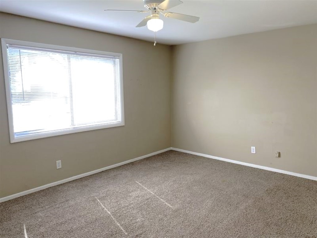 Empty room with beige walls and light brown carpet. A large window on the left allows natural light to fill the space, and a ceiling fan is visible above. The room appears clean and uncluttered, creating a bright and open atmosphere.