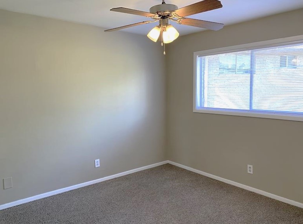 A minimally furnished room with beige walls and carpet. A ceiling fan with wooden blades is mounted in the center, and there is a large window with light filtering through, allowing natural light into the space.