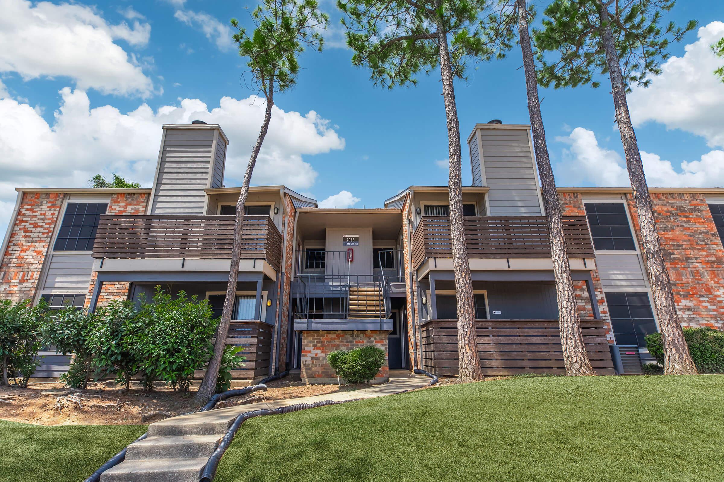 A two-story apartment building with brick and wooden exterior, featuring balconies on the upper level. Lush green landscaping surrounds the building, and tall pine trees are visible in the background against a bright blue sky with fluffy clouds. A walkway leads to the entrance.