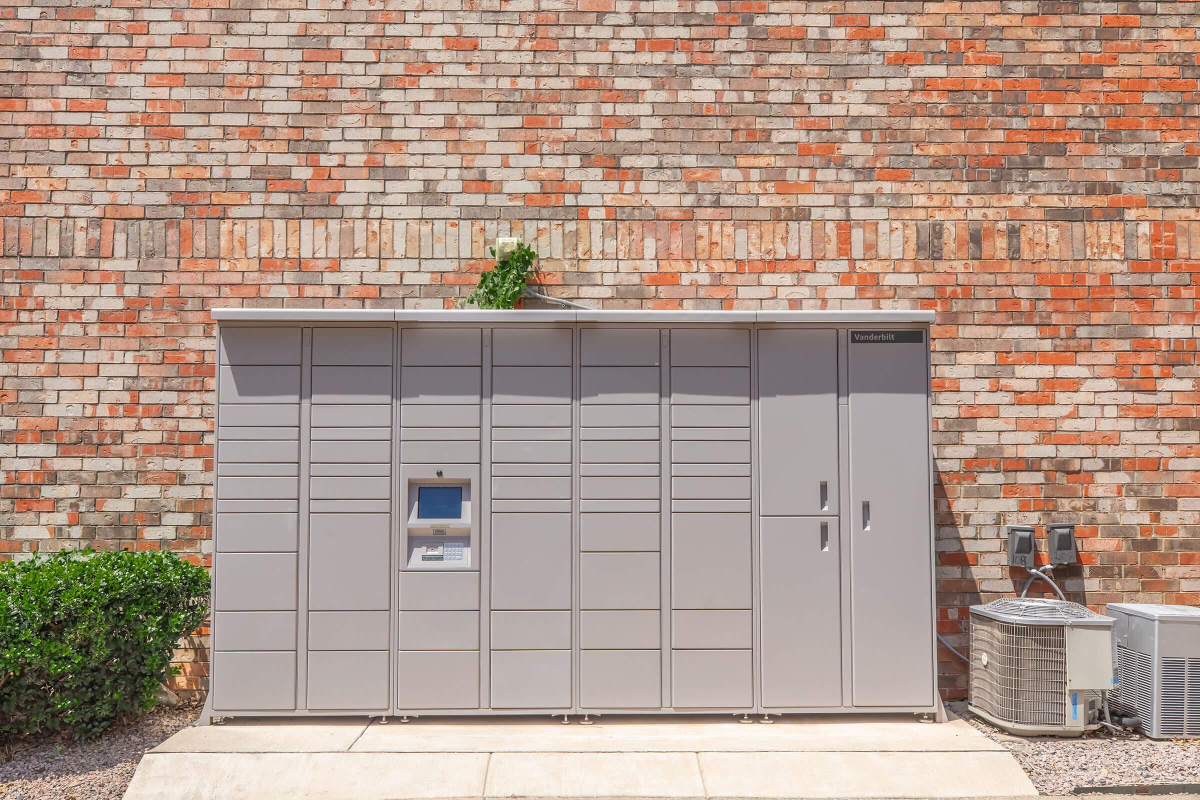 A large, gray package delivery locker against a brick wall, featuring a small touch screen and multiple compartments for parcel collection. Green plants are visible near the base, and air conditioning units are situated to the side. The setting is well-lit and organized, indicating a functional delivery area.