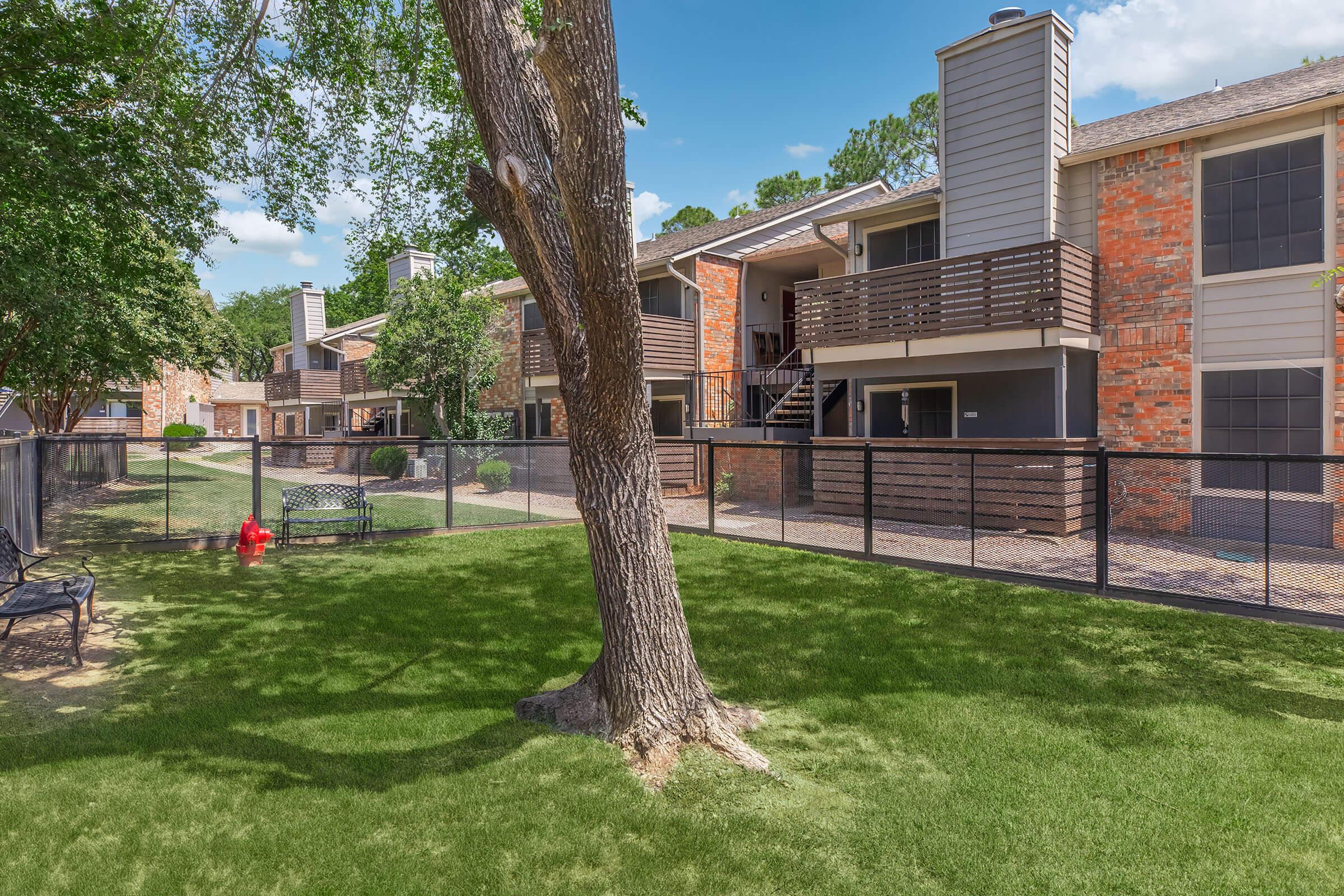 View of a landscaped courtyard in an apartment complex, featuring several brick buildings with balconies. A large tree stands in the foreground, and a black fence encloses the area. Green grass covers the ground, with a bench and fire hydrant visible in the yard under a clear blue sky.