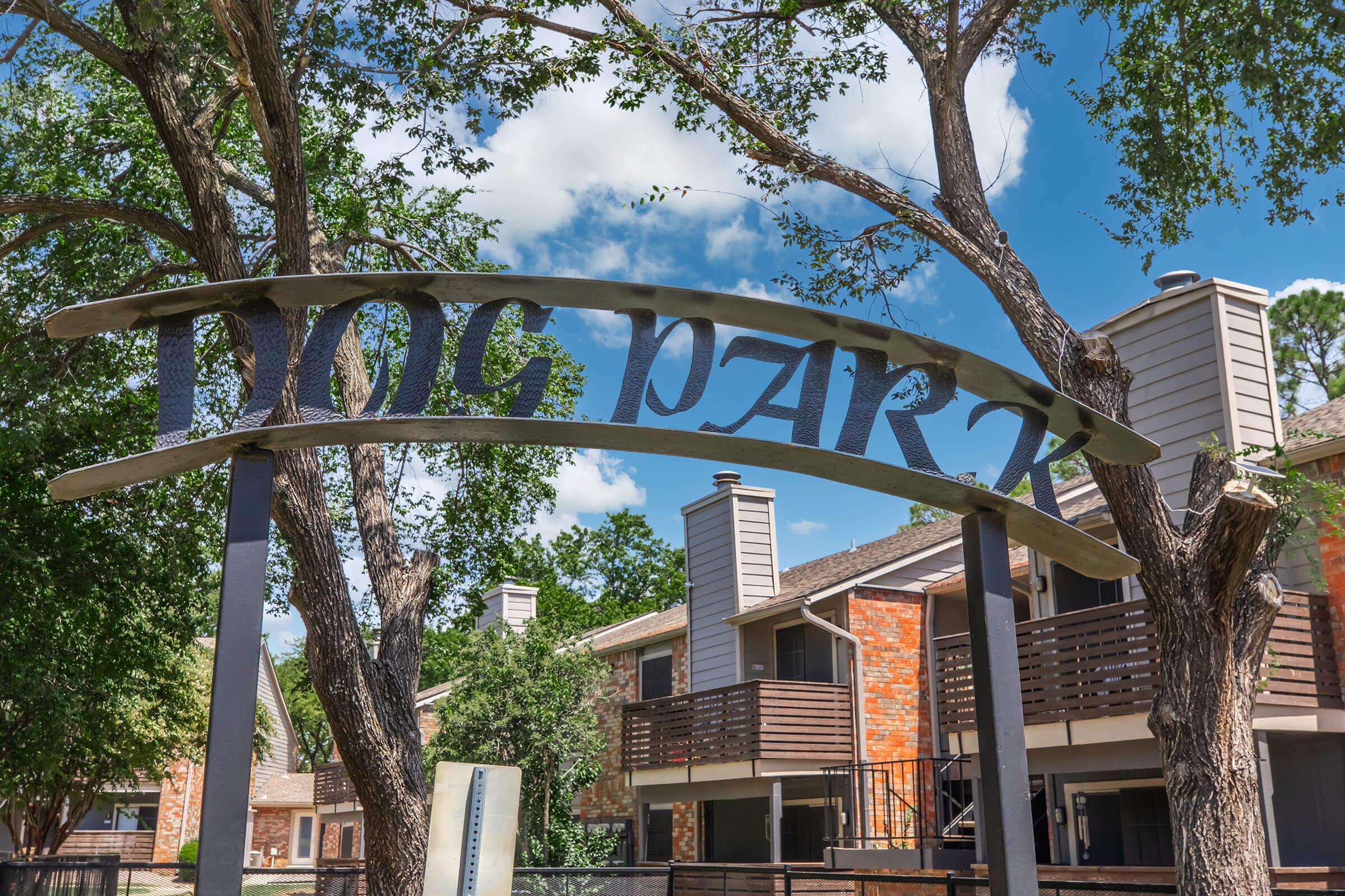 A sign that reads "DOG PARK" arching over a path, surrounded by trees and residential buildings in the background. The sky is bright with scattered clouds, creating a welcoming atmosphere for pet owners and their dogs.