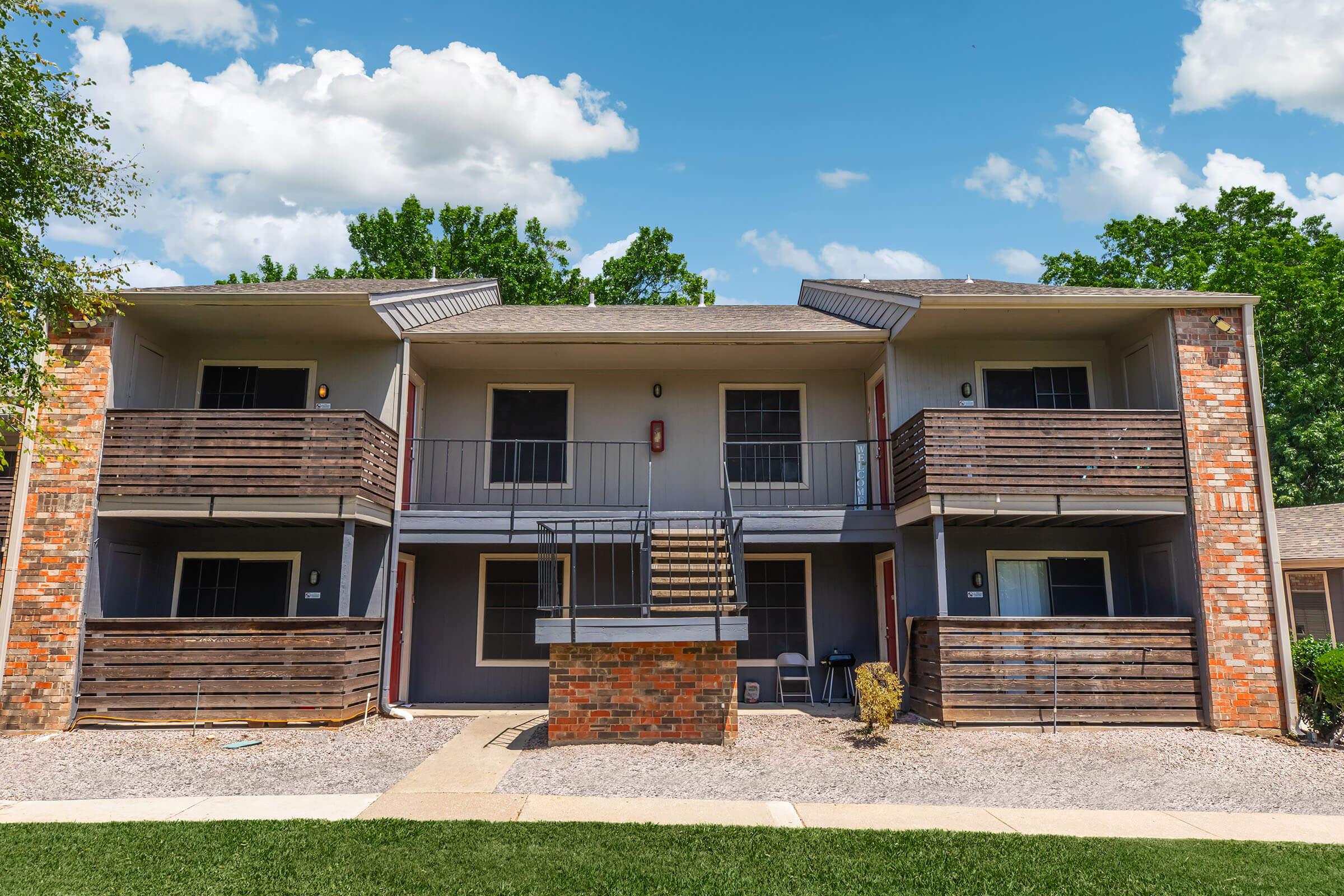 Two-story apartment building featuring a symmetrical design with wooden balconies on each side. The brick facade is complemented by light-colored walls and a gray roof. A concrete pathway leads to the entrance, surrounded by grassy areas and trees under a blue sky with clouds.