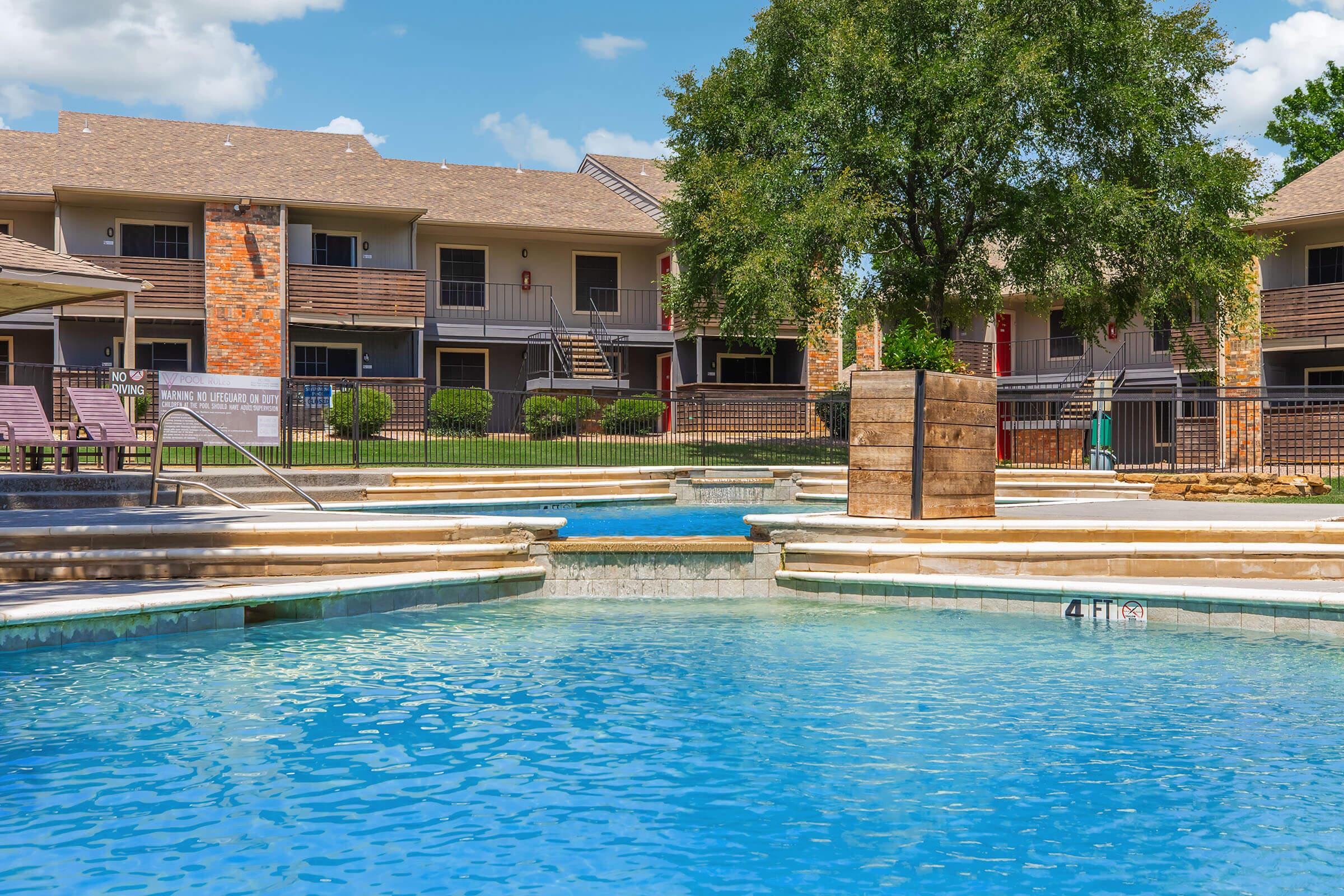 A swimming pool area surrounded by apartment buildings. The pool features a shallow section and a waterfall feature, with lounge chairs by the side. Lush greenery and a clear blue sky add to the relaxing atmosphere.