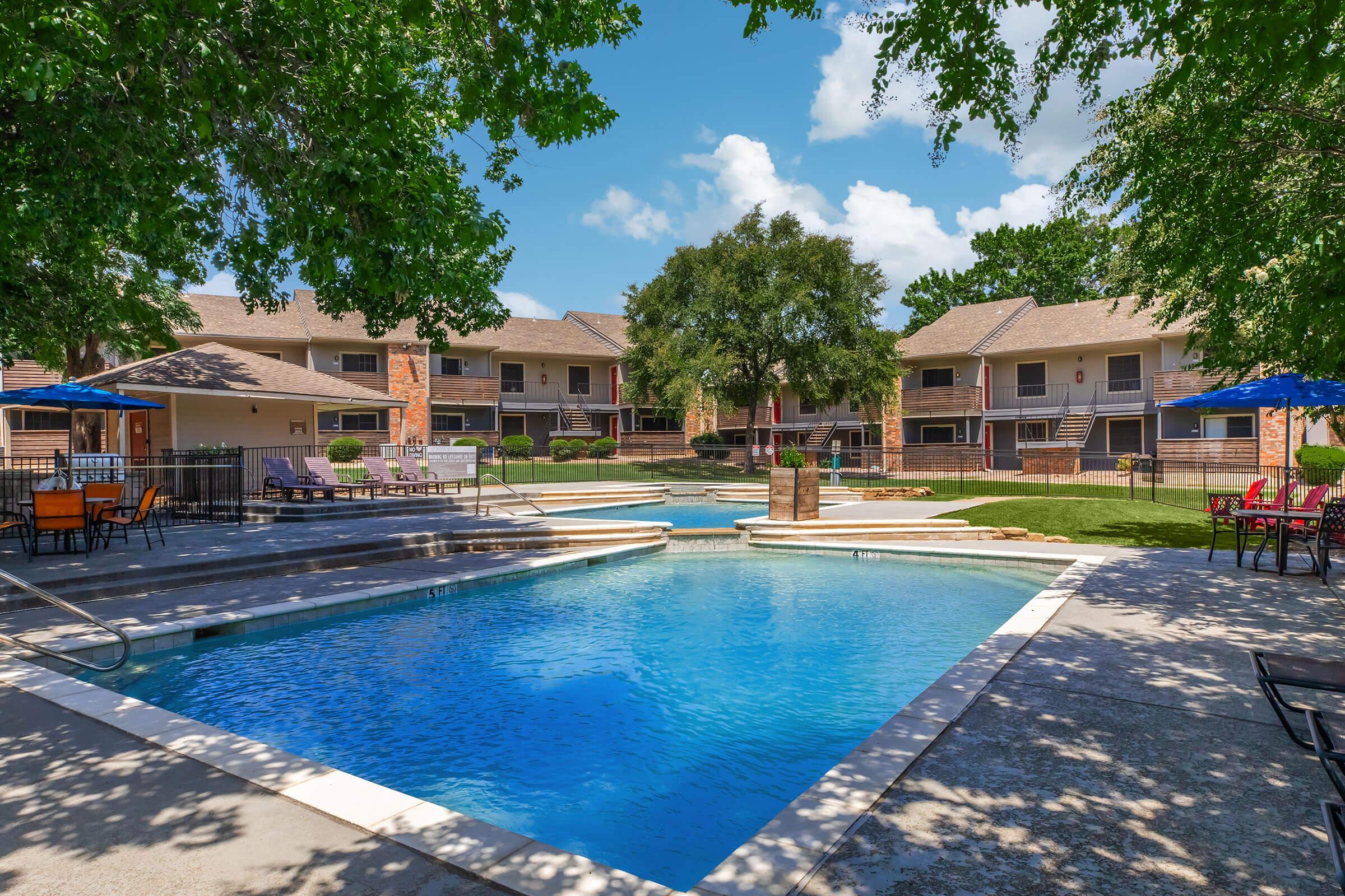 A tranquil outdoor pool area surrounded by residential buildings. The pool features a clear blue surface, with lounge chairs nearby and shade from trees. Well-maintained green lawns and walkways invite relaxation, complemented by bright blue skies and fluffy clouds above.