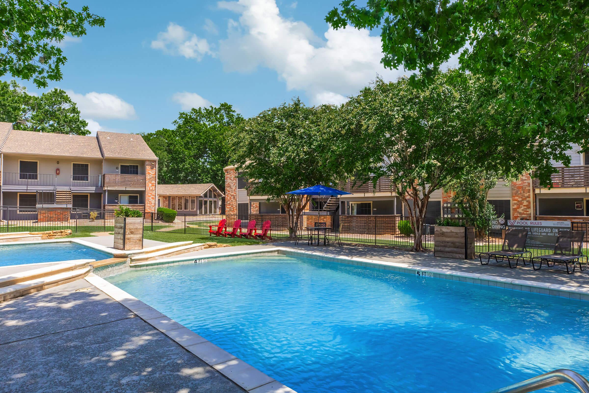 A view of a sunny outdoor pool area with two swimming pools, surrounded by lounge chairs and a shaded umbrella. In the background, there are brick apartment buildings and lush green trees, creating a relaxing environment. The sky is bright blue with a few fluffy clouds.