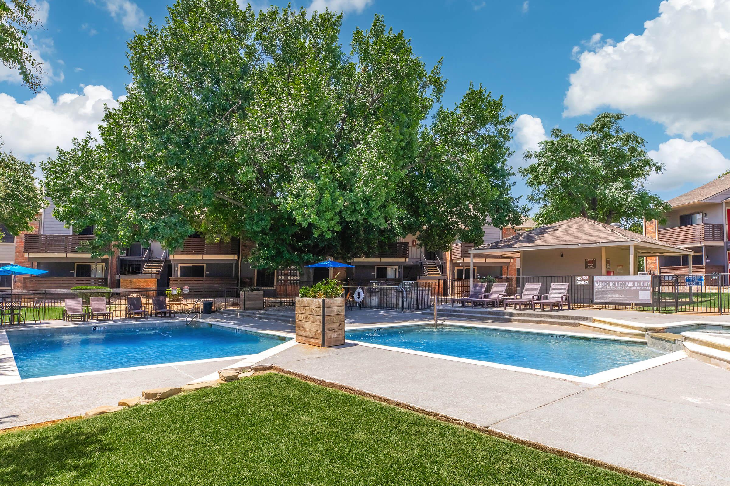 A scenic outdoor pool area featuring two connected swimming pools surrounded by lush green grass and lounge chairs. Large trees provide shade, and there are umbrellas near the pools. Behind the pools, there are residential buildings, creating a relaxed atmosphere on a sunny day.