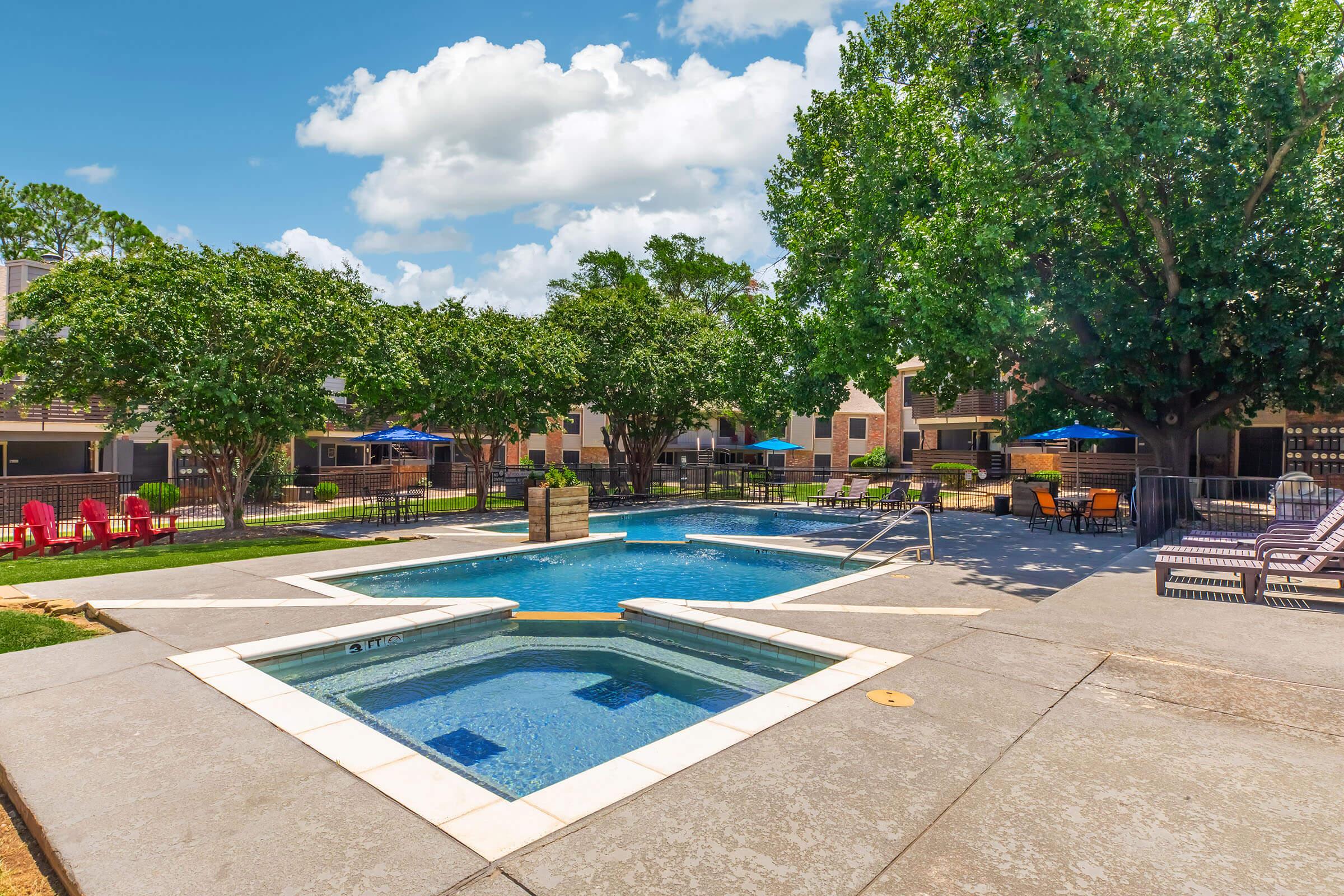 A sunny outdoor pool area featuring a swimming pool and a hot tub surrounded by greenery. Lounge chairs and red Adirondack chairs are positioned around the pool. Umbrellas provide shade, and apartment buildings can be seen in the background under a bright blue sky with fluffy white clouds.