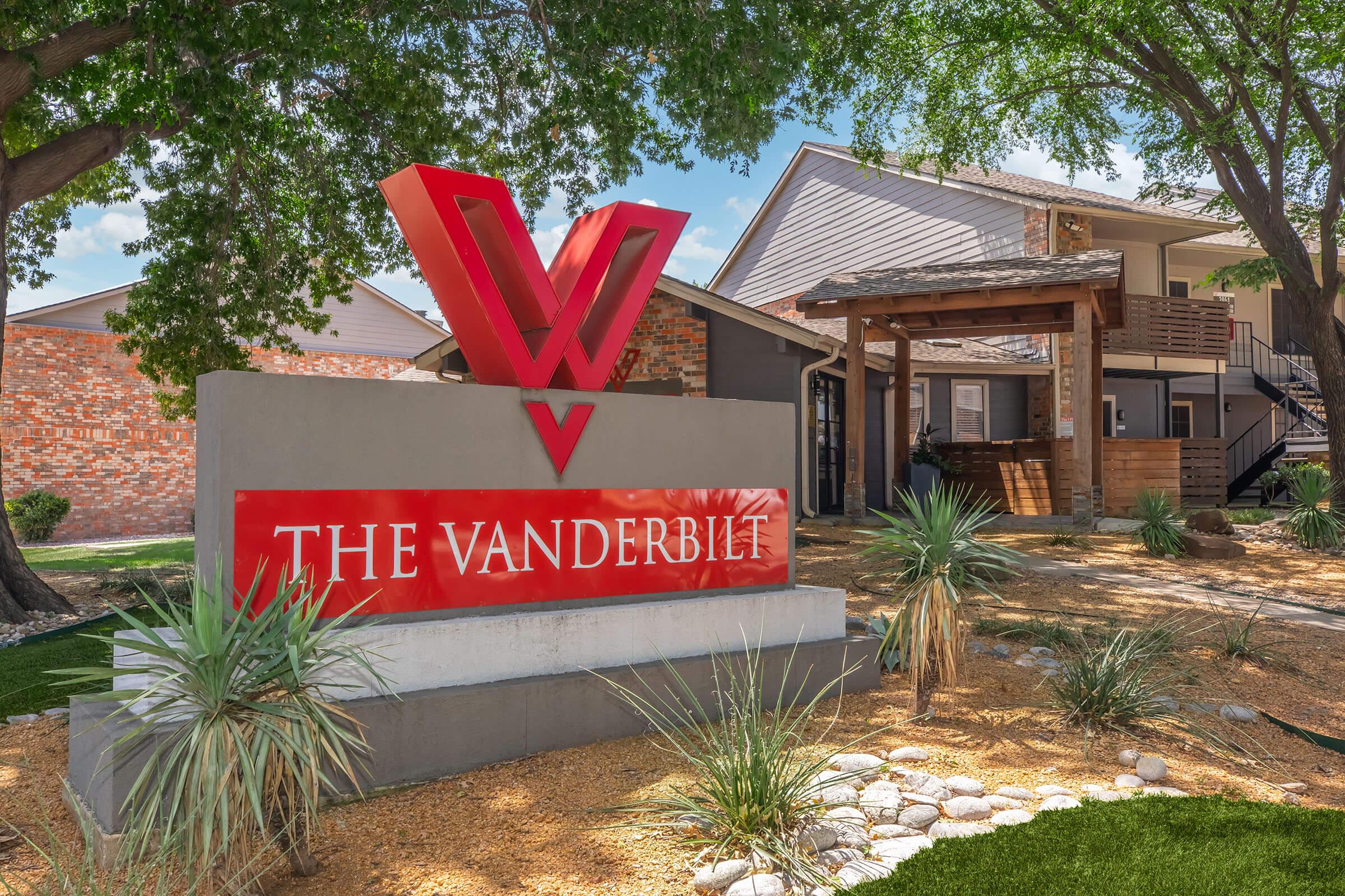 Sign for "The Vanderbilt" in bold red letters, with a stylized 'V' logo, set against a landscaped area featuring decorative rocks and plants. In the background, a modern apartment building is partially visible, surrounded by trees and greenery.