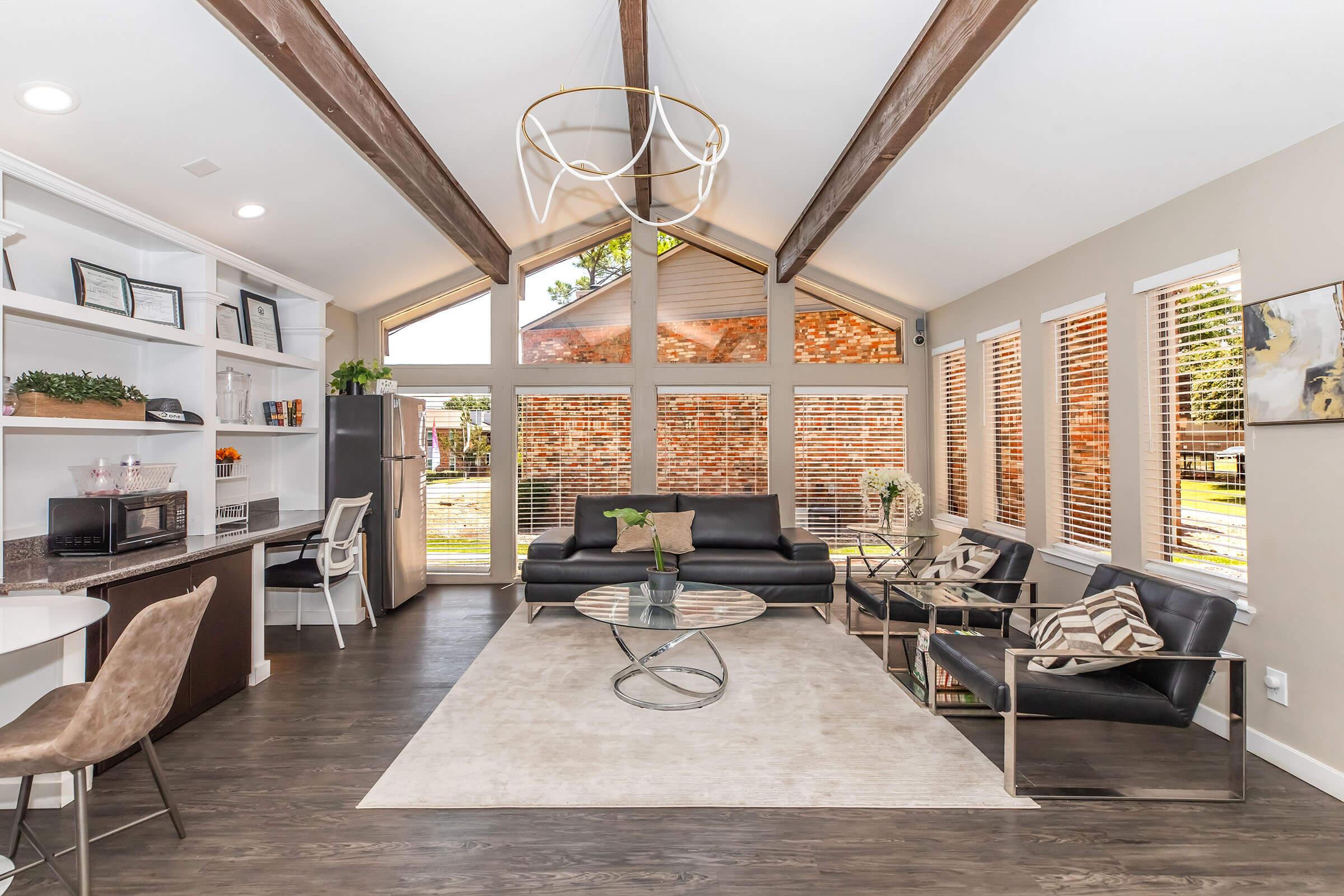 A modern living room with vaulted ceilings, wooden beams, and large windows letting in natural light. The space features a black sofa, glass coffee table, stylish chairs, and built-in shelves with decorative items. The floor is dark wood, and there are neutral-colored accents throughout, creating a bright and inviting atmosphere.