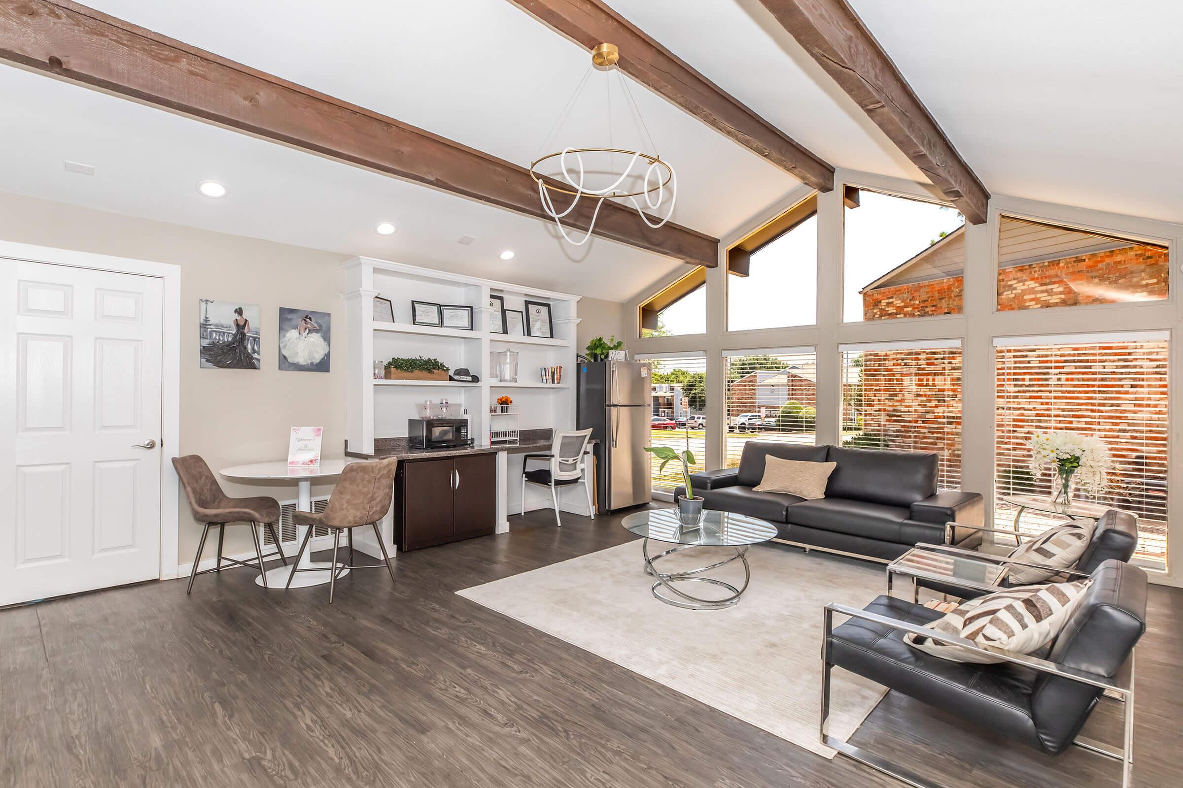 A modern living room featuring high ceilings with exposed wooden beams, large windows allowing natural light, a sleek black sofa, stylish armchairs, a coffee table, and a bookshelf with decorative elements. The space has a warm, inviting atmosphere with neutral tones and contemporary decor.