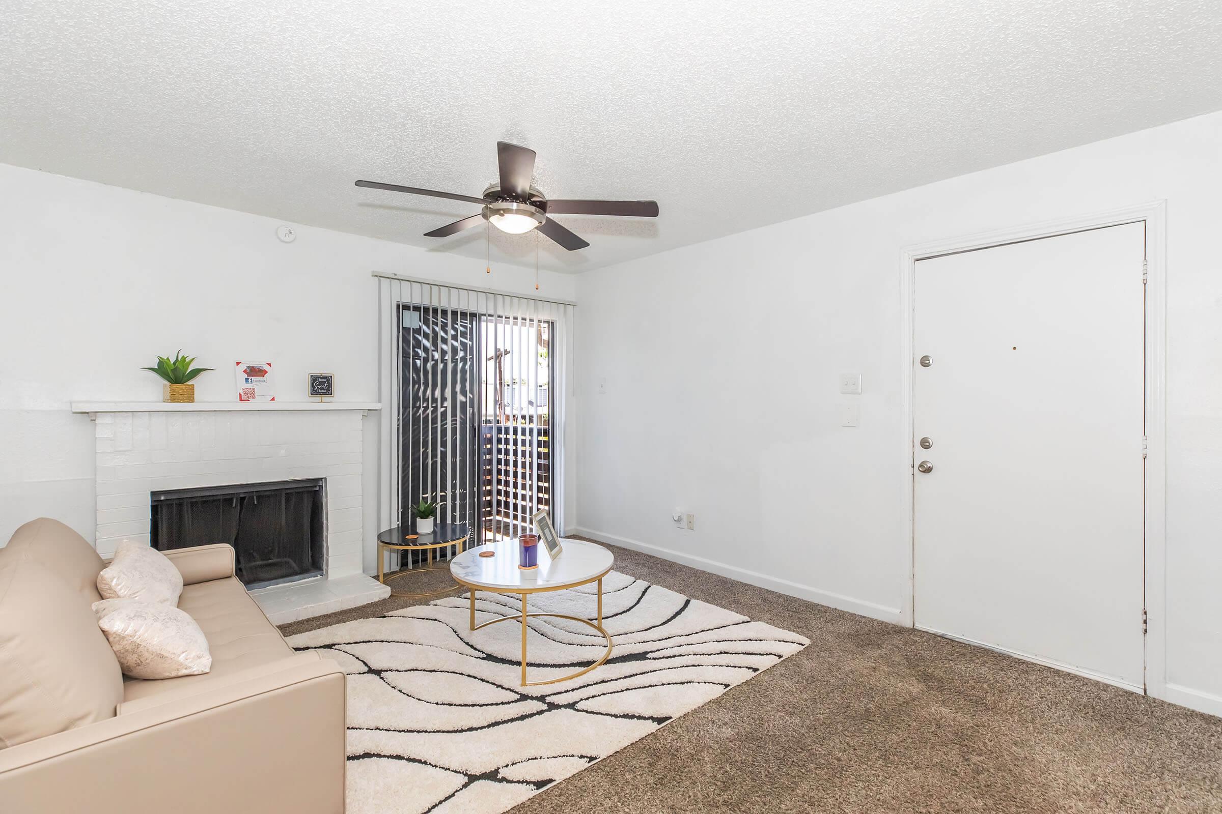 A cozy living room featuring a beige sofa, a round coffee table, and a decorative area rug. There's a fireplace with a small plant above it, and a glass door leading to a balcony. The walls are painted white, and a ceiling fan is visible. The space is well-lit with natural light.