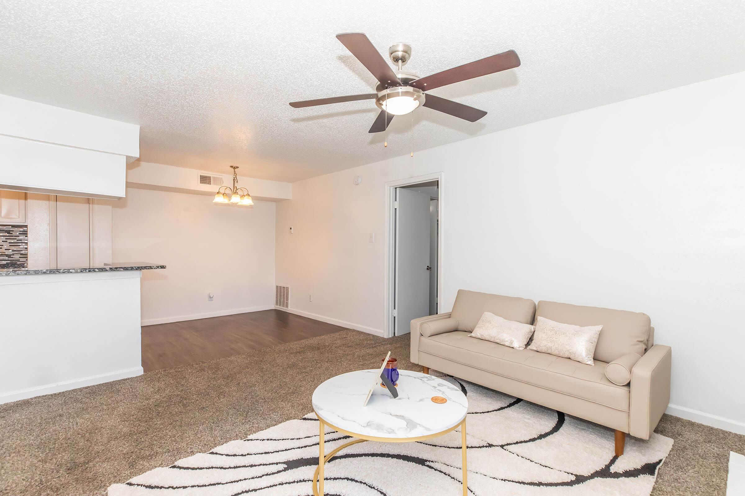 A cozy living room featuring a light-colored sofa, a round coffee table with decor, and a ceiling fan. The space has neutral wall colors, plush carpeting, and a small dining area with pendant lighting in the background, along with an open doorway leading to a different room.