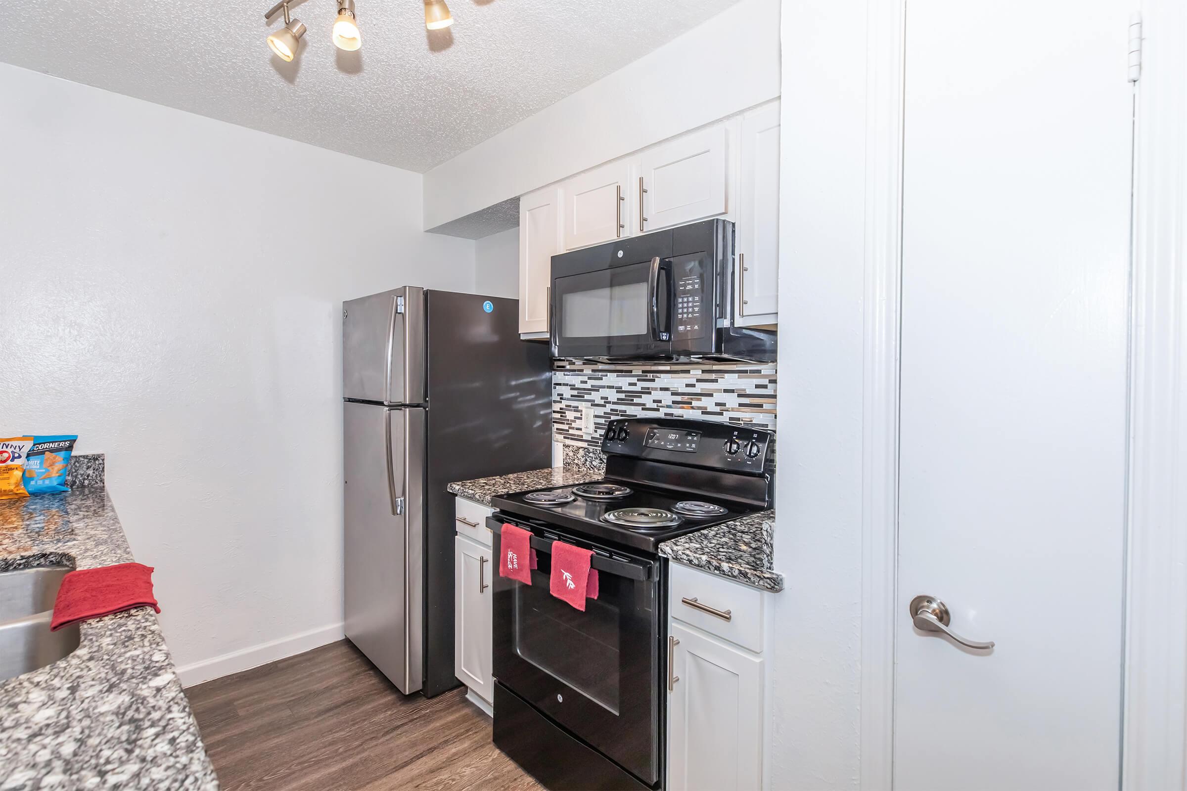 Modern kitchen with stainless steel appliances including a refrigerator and oven. The countertops are dark granite, and there's a decorative backsplash. Two red towels hang from the stove, and the lighting is bright. The floor has a wood-like finish, creating a stylish and functional cooking space.