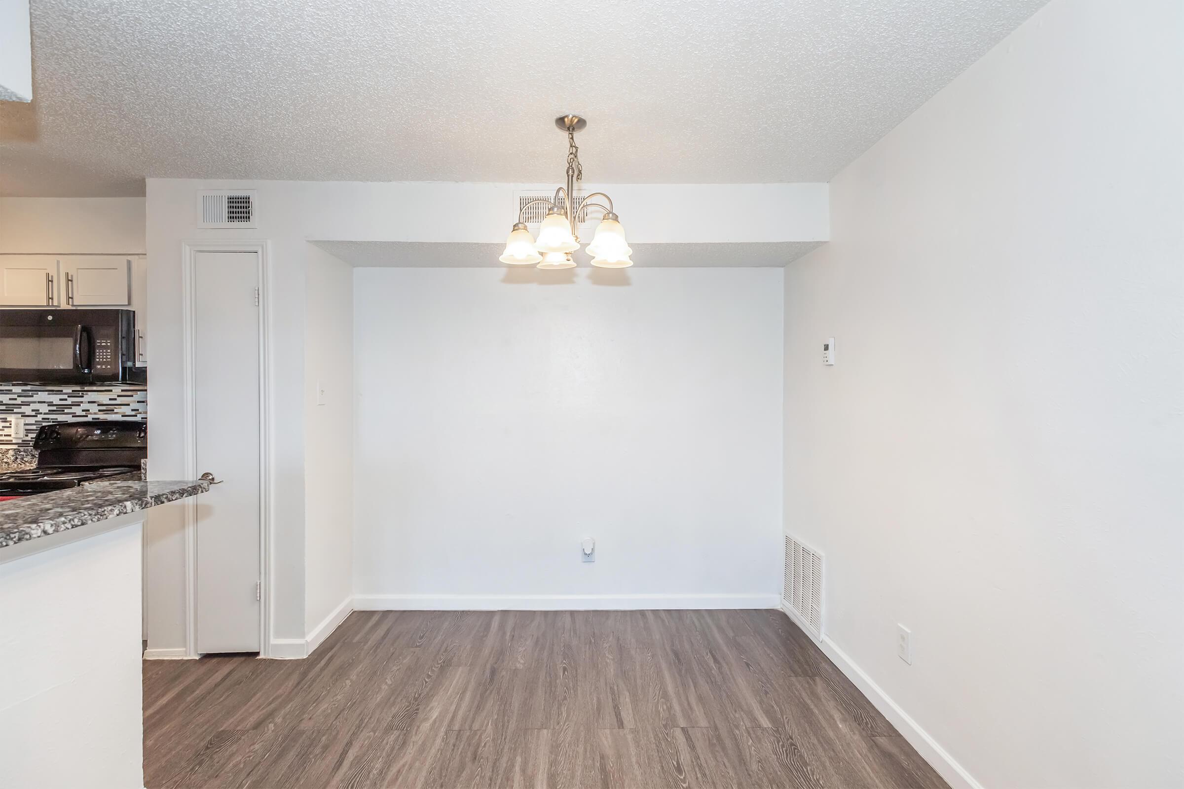 A well-lit dining area with a modern chandelier and hardwood-style flooring. The space features white walls and a small wall-mounted appliance. To the left, there is an opening leading to a kitchen with granite countertops and dark cabinetry. The area is clean and uncluttered, creating a spacious feel.