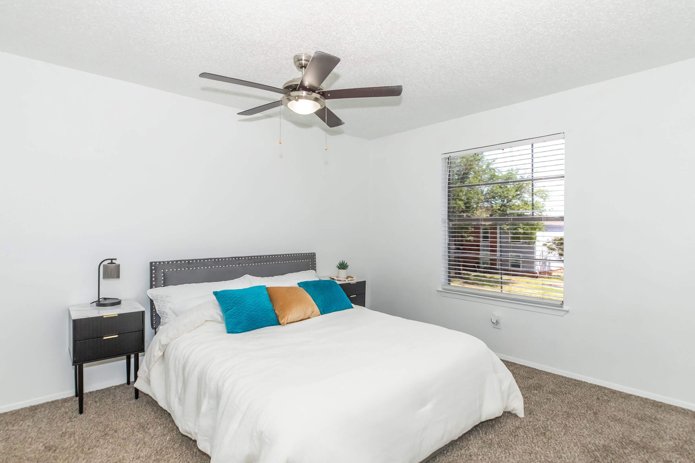 A bright and airy bedroom featuring a queen-sized bed with white bedding and decorative blue and gold pillows, a modern black nightstand with a lamp, a ceiling fan, and a window with blinds that allows natural light. The walls are painted white and the flooring is carpeted.