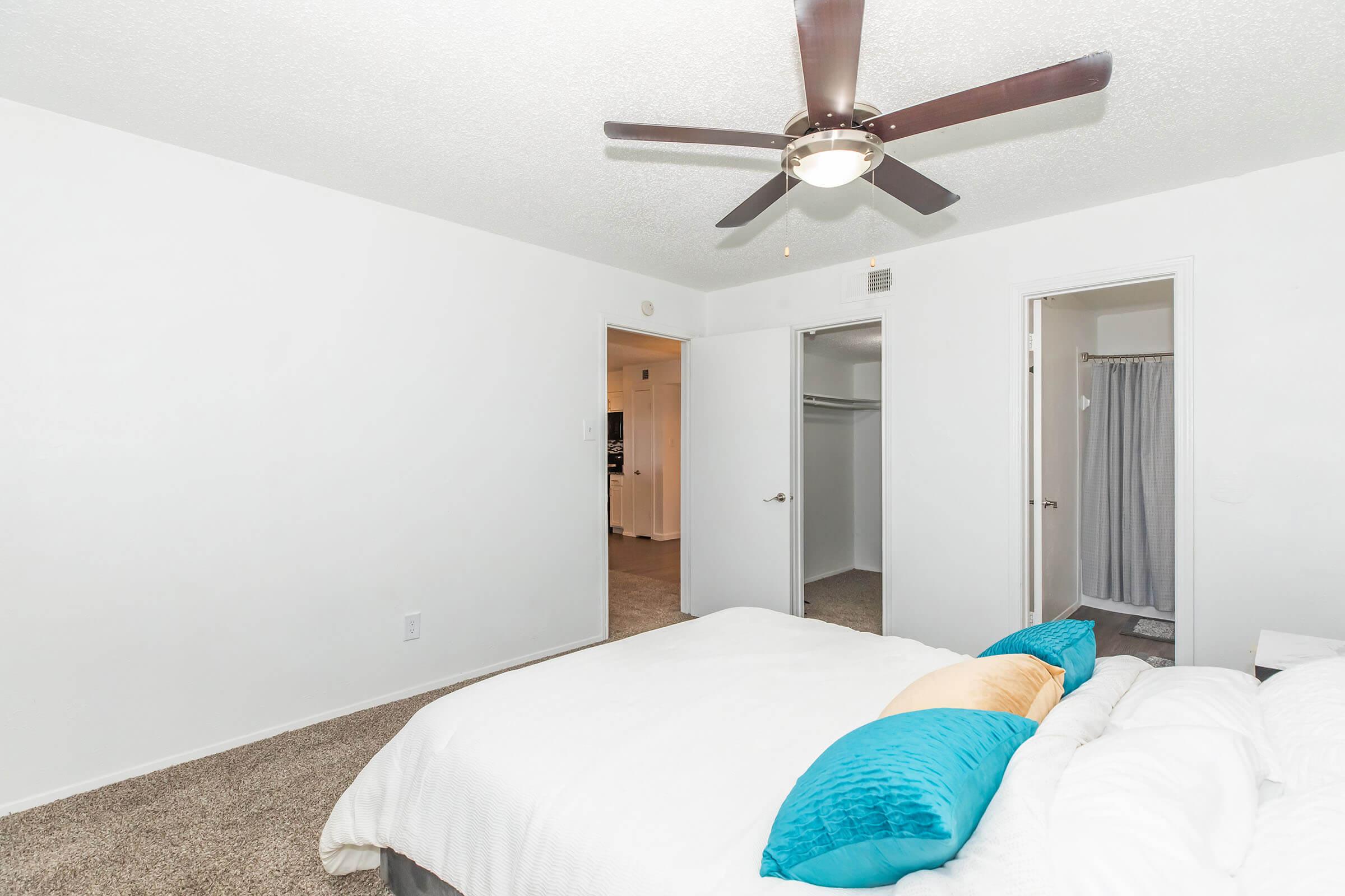 A modern bedroom featuring a large bed with white bedding and decorative pillows in shades of blue and orange. There is a ceiling fan above, light-colored walls, and carpeted flooring. Two open doorways lead to closets and a bathroom area, providing a bright and spacious feel.