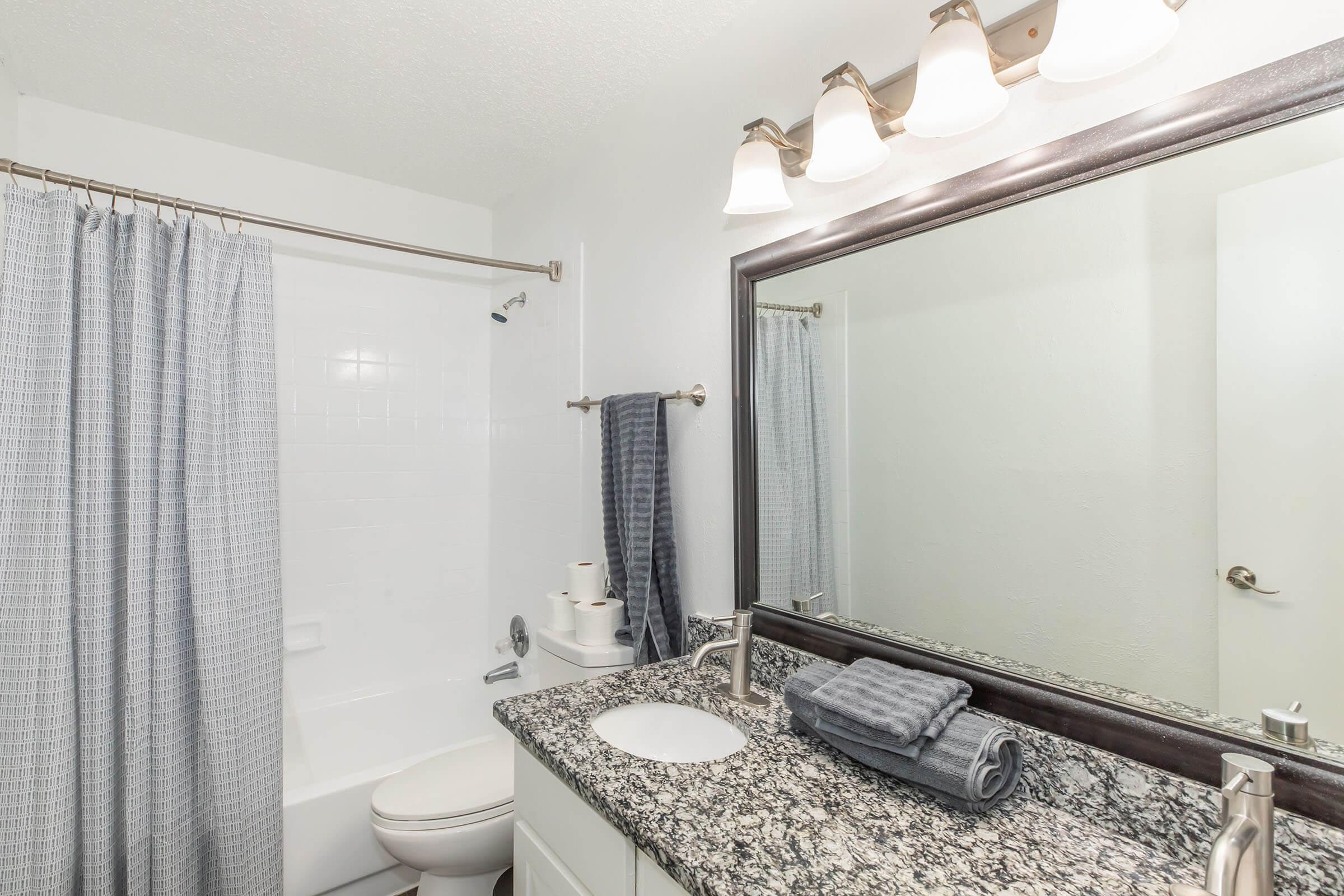 A modern bathroom featuring a white bathtub, a gray shower curtain, a large mirror above a granite countertop, and stylish lighting. Neatly arranged towels in gray tones are placed on the counter, enhancing the clean and minimalist aesthetic. The walls are painted white, contributing to a bright ambiance.