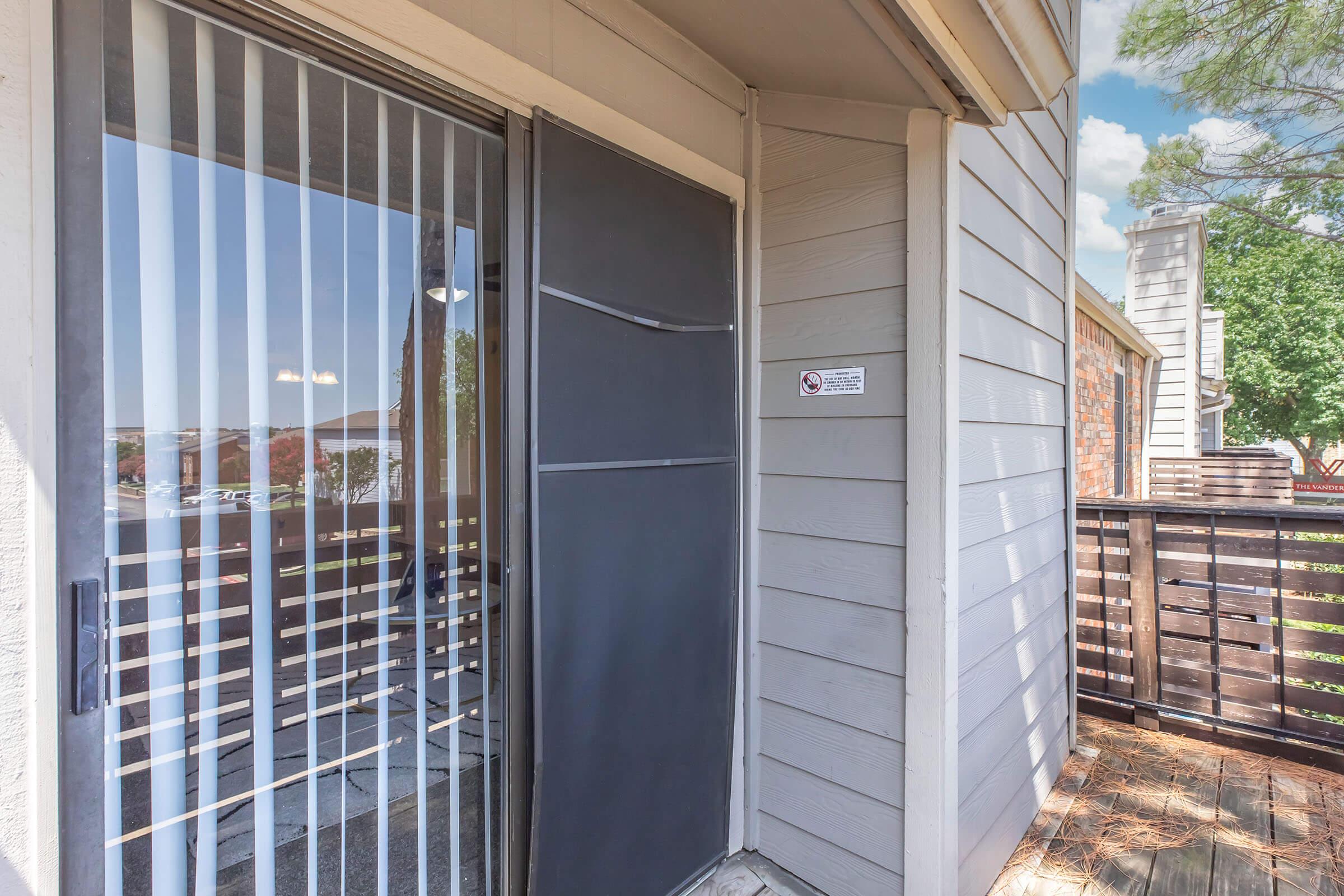 A covered entrance with a black security door, featuring vertical blinds and a sign. The porch has a wooden railing, and a glimpse of greenery is visible in the background, suggesting a residential area. The scene is well-lit and inviting.