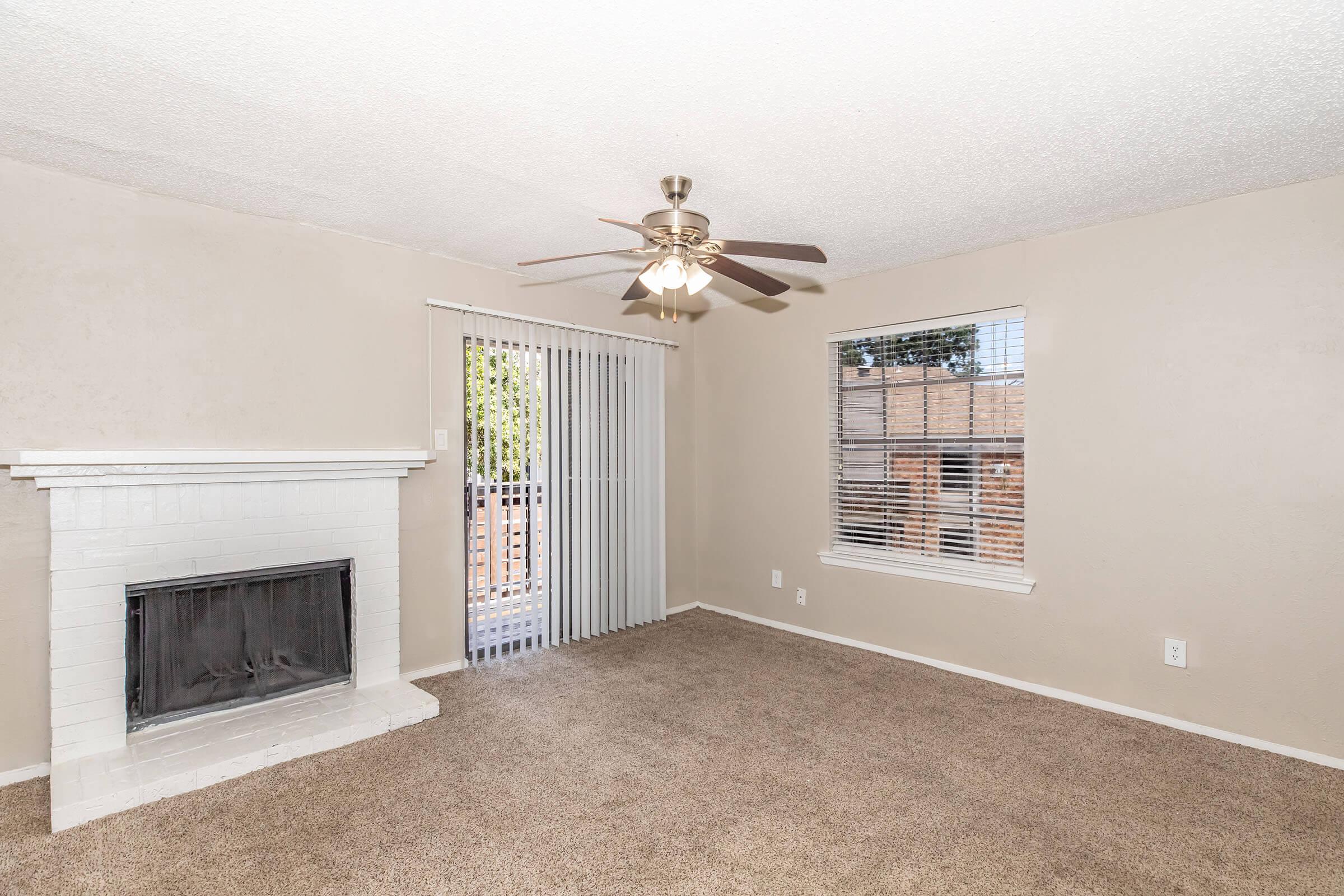 A cozy living room featuring a ceiling fan, a white brick fireplace, and large windows with blinds. The room has beige walls and plush carpet, creating a warm atmosphere. A sliding glass door leads outside, allowing natural light to fill the space.
