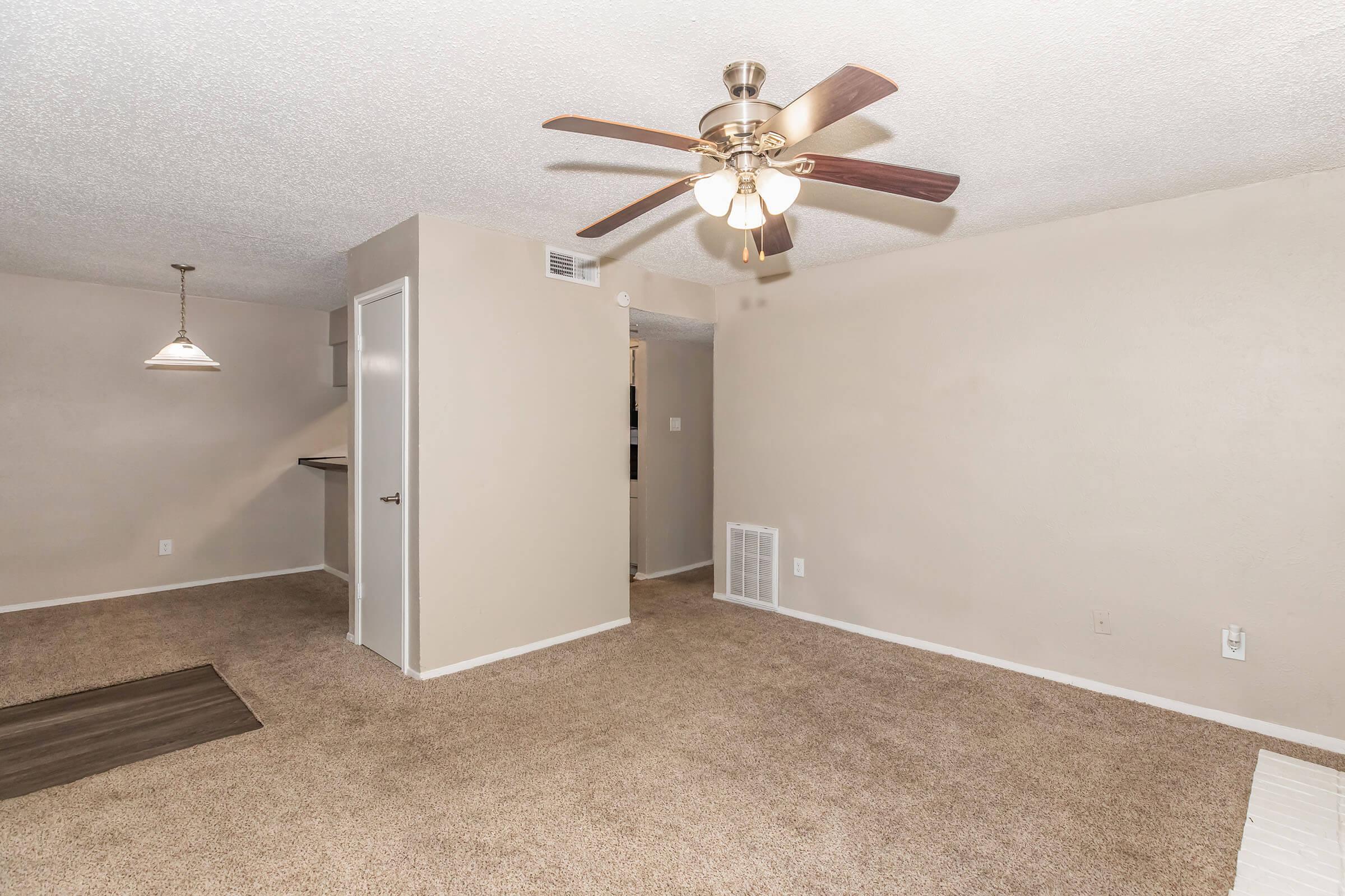 A spacious, well-lit living room featuring a ceiling fan, beige walls, and carpeted flooring. There is a doorway leading to another room and a small kitchenette visible in the background. A pendant light hangs from the ceiling, adding a cozy atmosphere to the space.