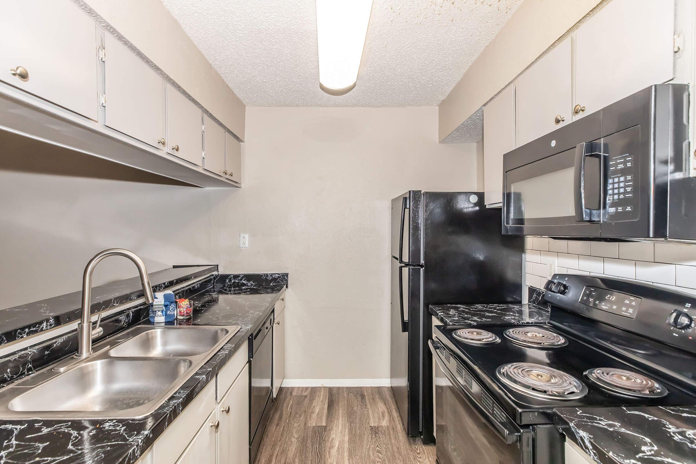 Modern kitchen featuring black appliances, including a refrigerator, microwave, and stove. The countertops have a black marble finish, and light-colored cabinets line the walls. There is a dual sink with a faucet and ample cabinet space above, set against a neutral-colored wall. Natural light is minimized by the overhead light.