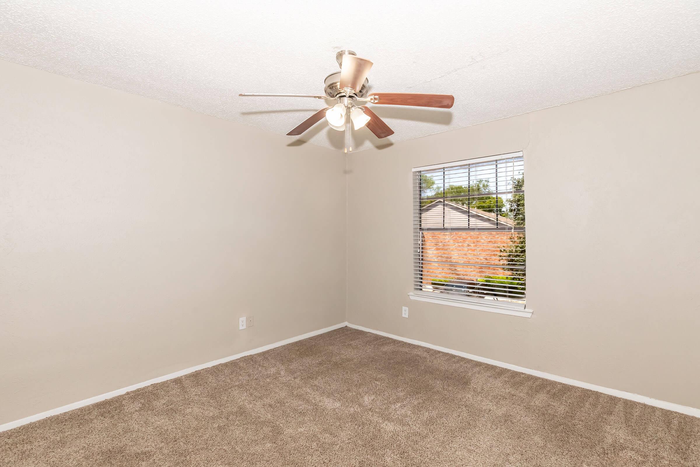 Empty room with light beige walls and plush carpet. A ceiling fan with wooden blades hangs in the center. There's a window with blinds on the right, allowing natural light in and revealing a brick exterior outside. The room appears spacious and clean, ready for furnishing.