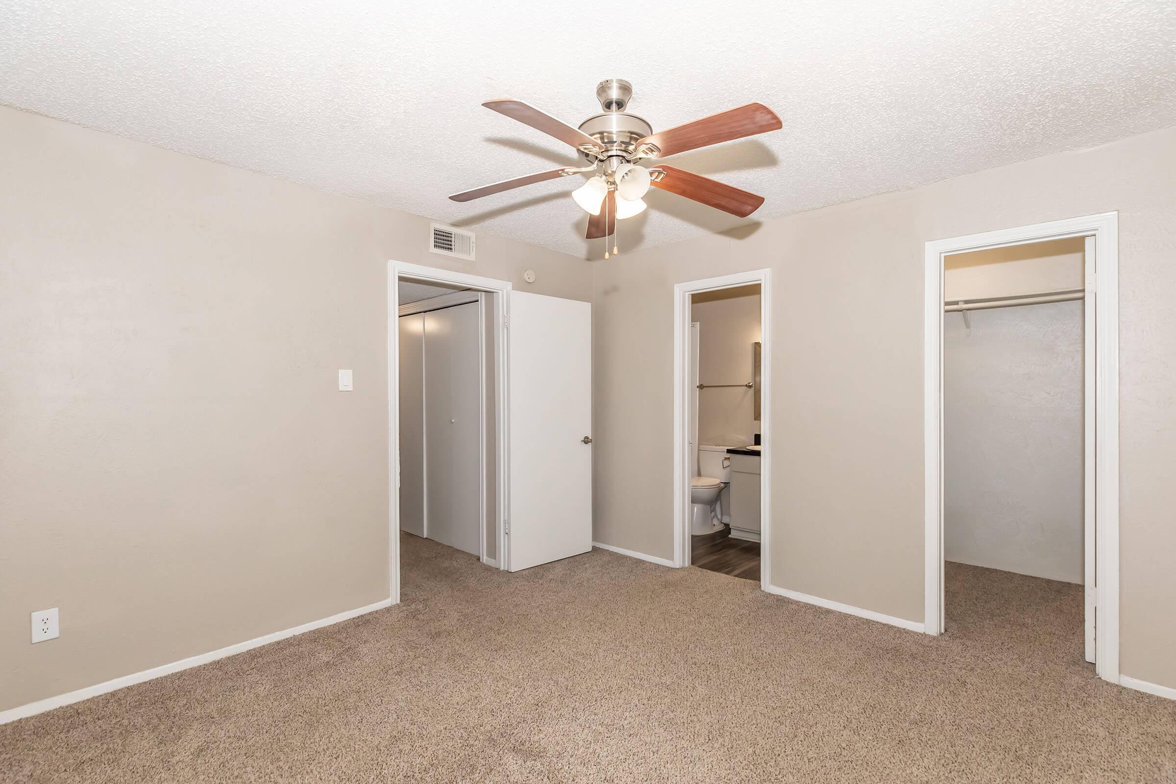 A spacious room with light brown carpet, featuring a ceiling fan. There are two doorways leading to a bathroom and a closet on the right. The walls are a light beige color, and the ceiling has a textured finish. The room is well-lit and has a clean, contemporary look.