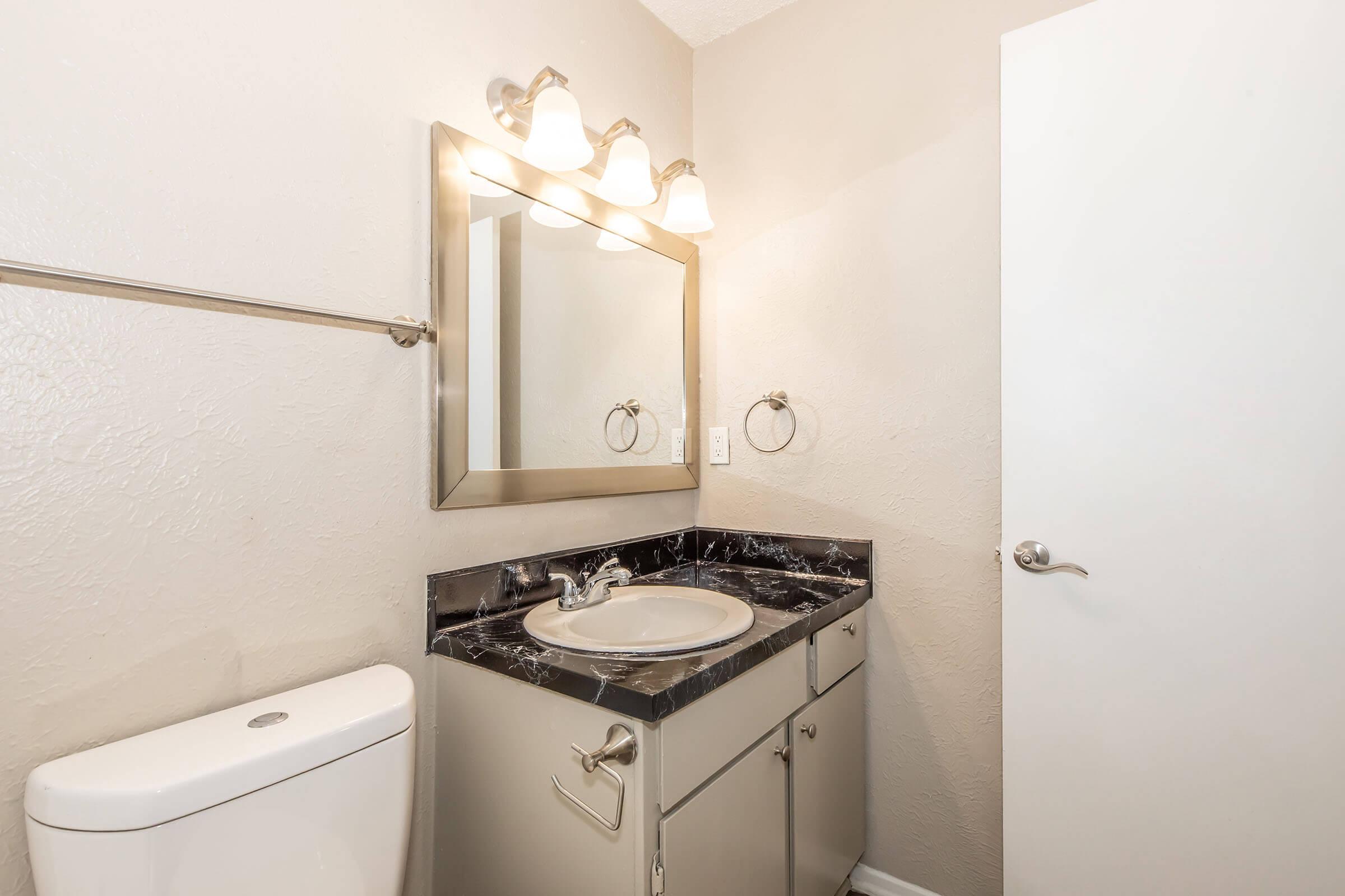 A modern bathroom featuring a white toilet, a sleek vanity with a black marble countertop, and a round sink. Above the vanity, there is a large mirror framed in brushed metal, along with three light fixtures. The walls are painted in a soft beige color, and a door is visible on the right side of the image.