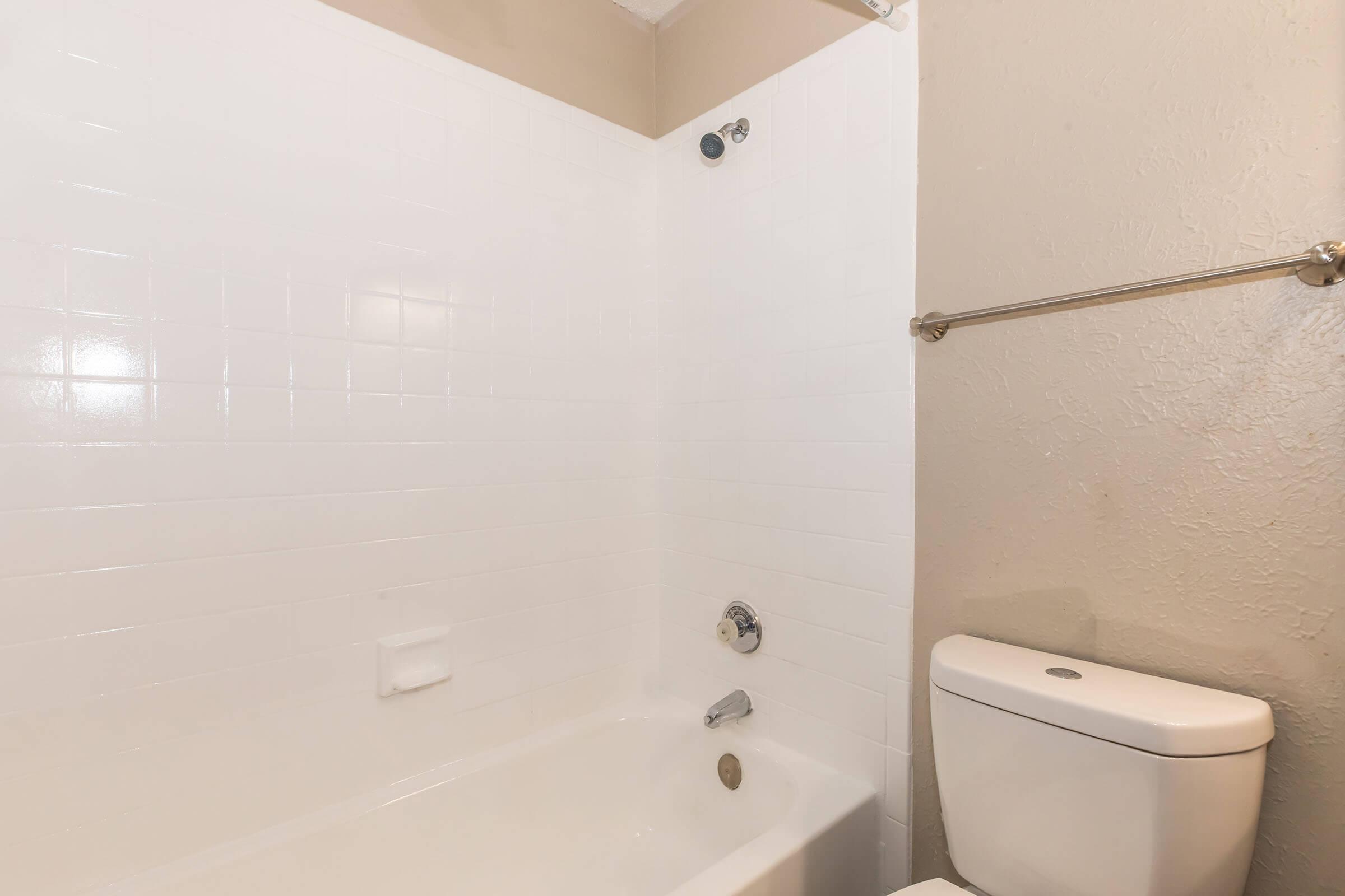 A clean, simple bathroom featuring a bathtub with a showerhead, white tiled walls, and a toilet. The space is well-lit, with neutral-colored walls and minimal fixtures, conveying a straightforward and functional design.
