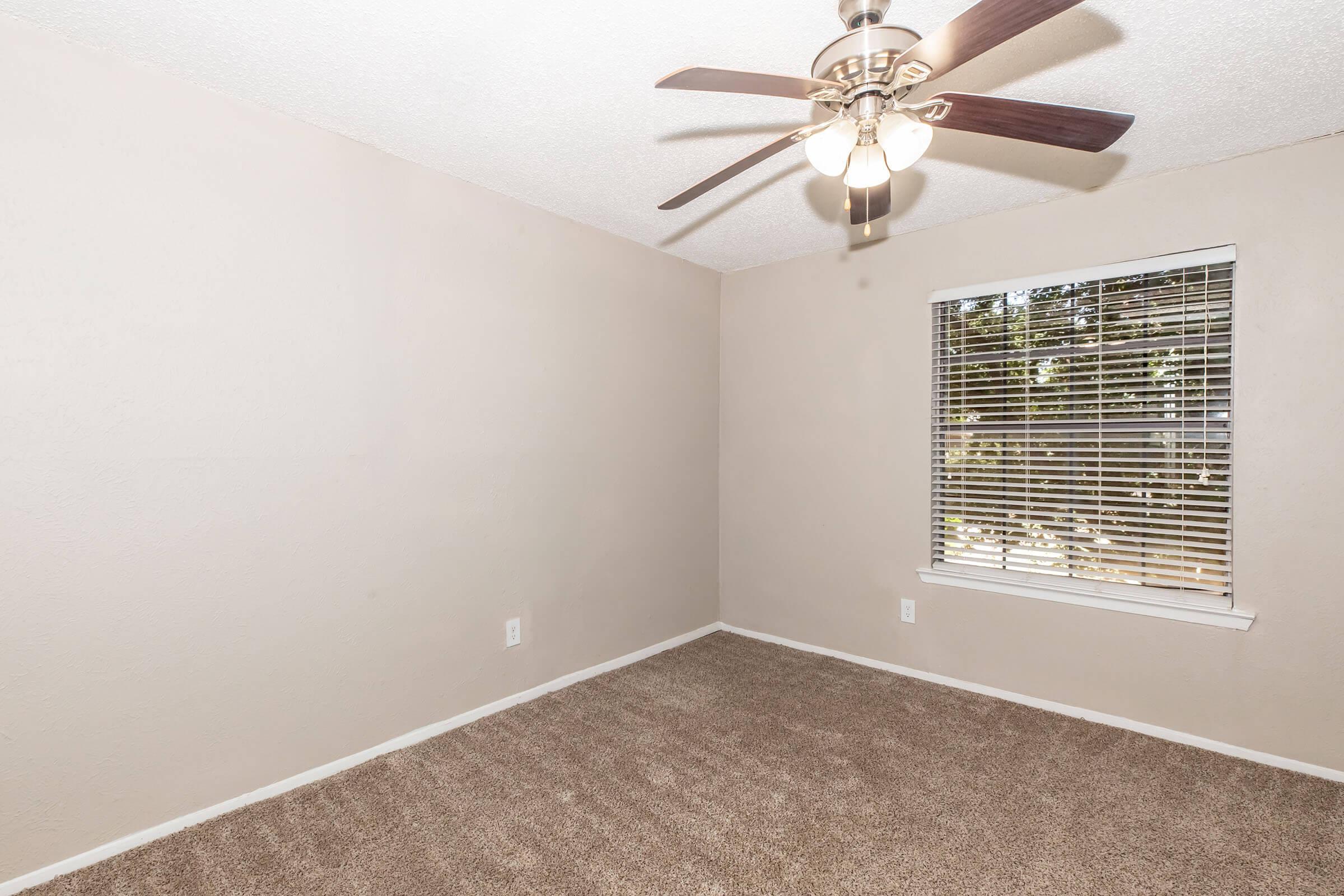A small, empty bedroom with light brown walls and carpet. A ceiling fan with wooden blades hangs above. A window with blinds allows natural light to enter, showcasing greenery outside. The room has a clean, uncluttered appearance, ideal for personalization or movement in.