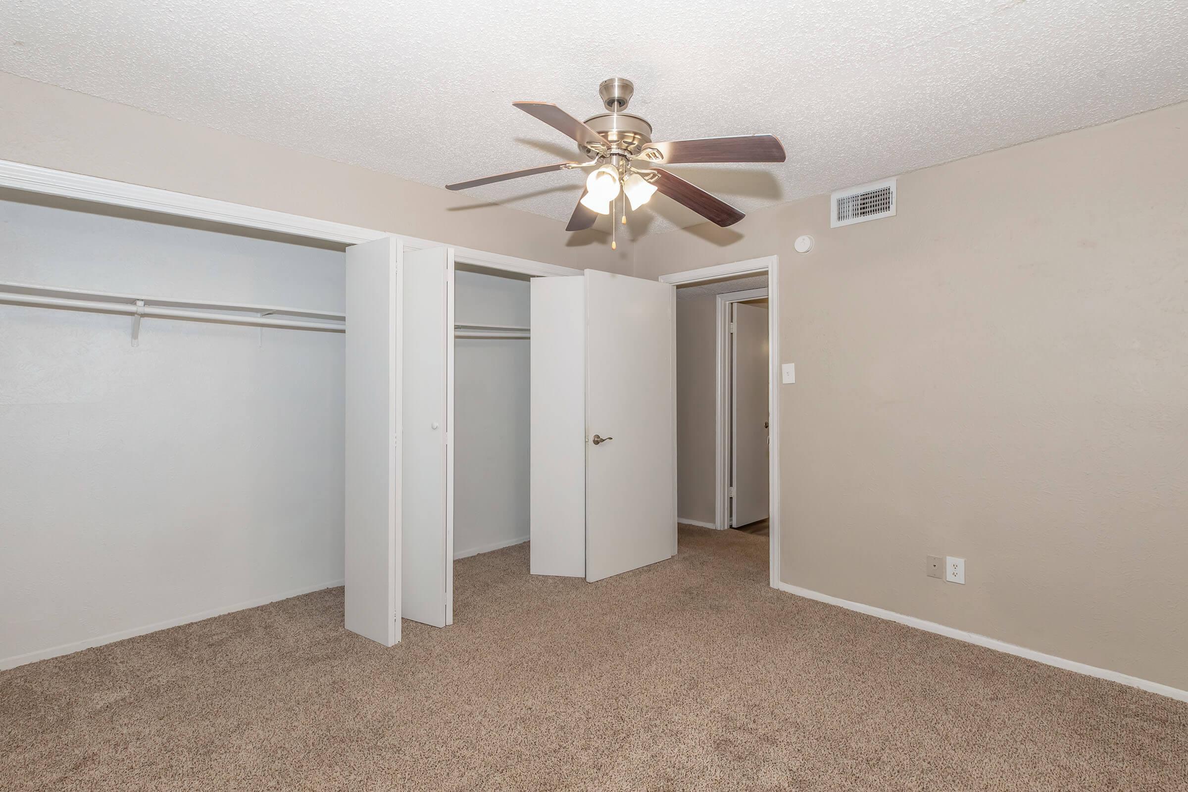 Empty bedroom with beige walls and carpet. A ceiling fan with light fixtures hangs from the ceiling. Two white double doors open to a closet on one side, while another door leads to an adjacent room. Natural light enters from a wall vent. Simple, clean decor.