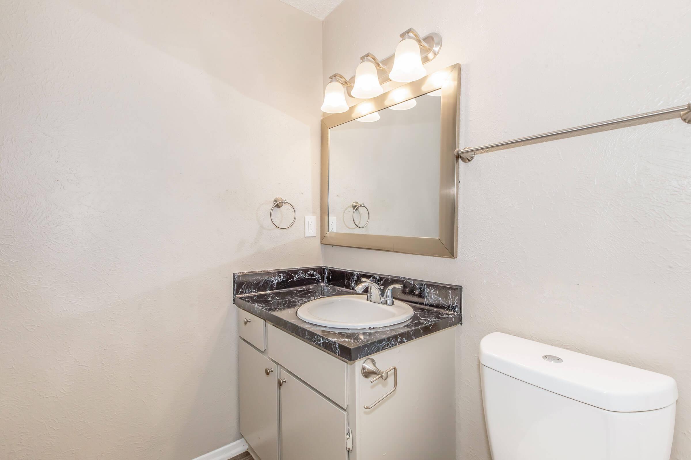 A well-lit bathroom featuring a granite countertop sink, a large mirror with three light fixtures above, a towel ring, and a modern toilet. The walls are painted in a light color, and the flooring appears to be laminate. The overall design is sleek and contemporary.