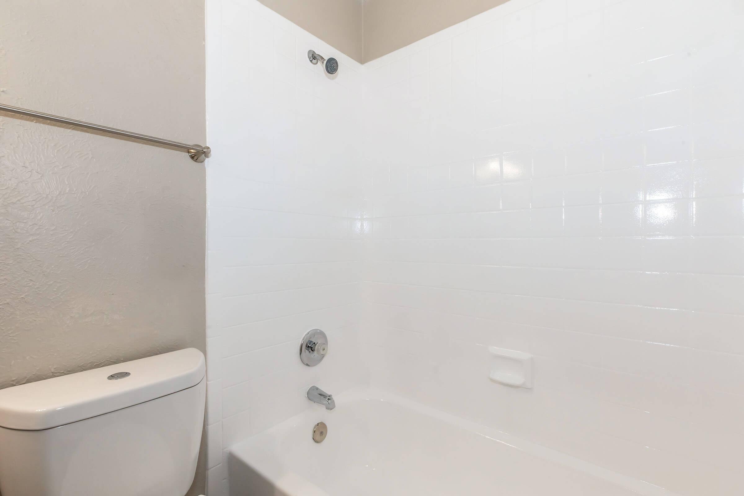 A clean, white bathroom featuring a bathtub with chrome fixtures, a showerhead, and a toilet. The walls are tiled in white, creating a bright and simple environment. A towel bar is mounted on the wall, and the space appears well-maintained.