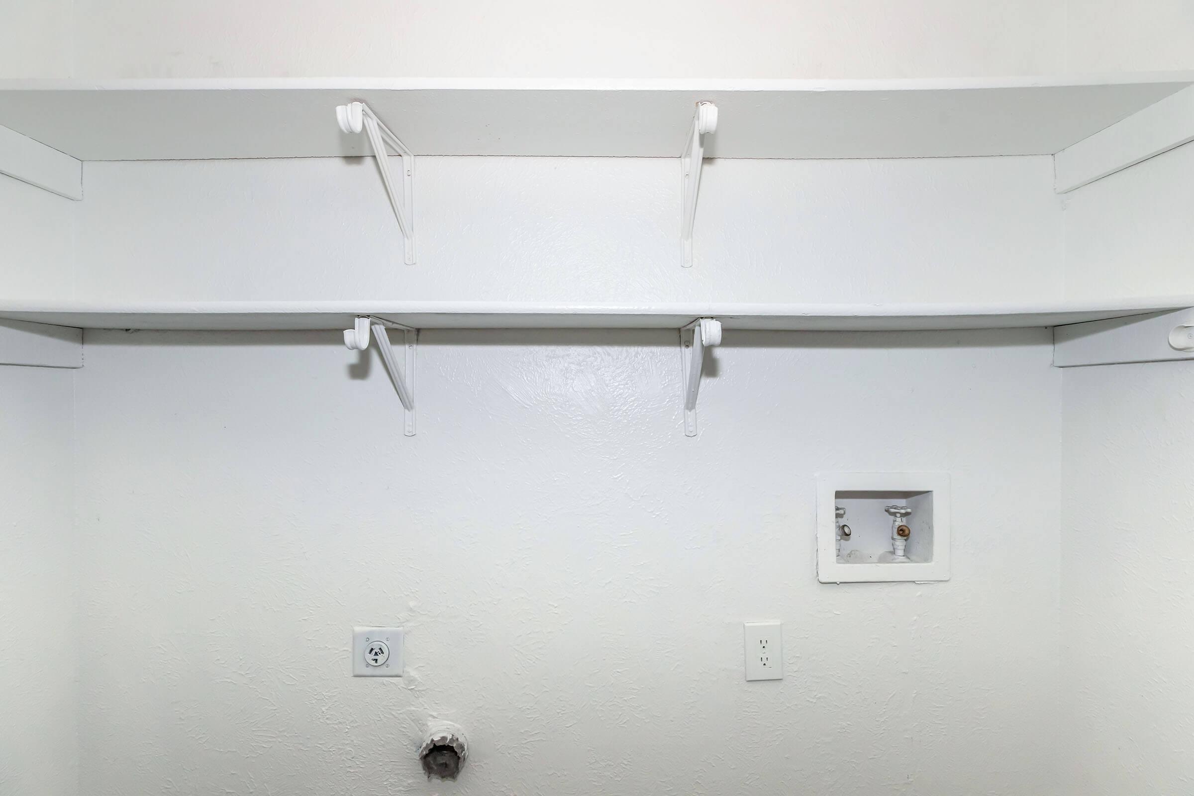 Empty white closet with two upper shelves and a small panel on the wall. There are electrical outlets and plumbing fixtures visible, but the space is mostly bare and unoccupied. The walls and shelves are painted white, giving a clean and simple appearance.
