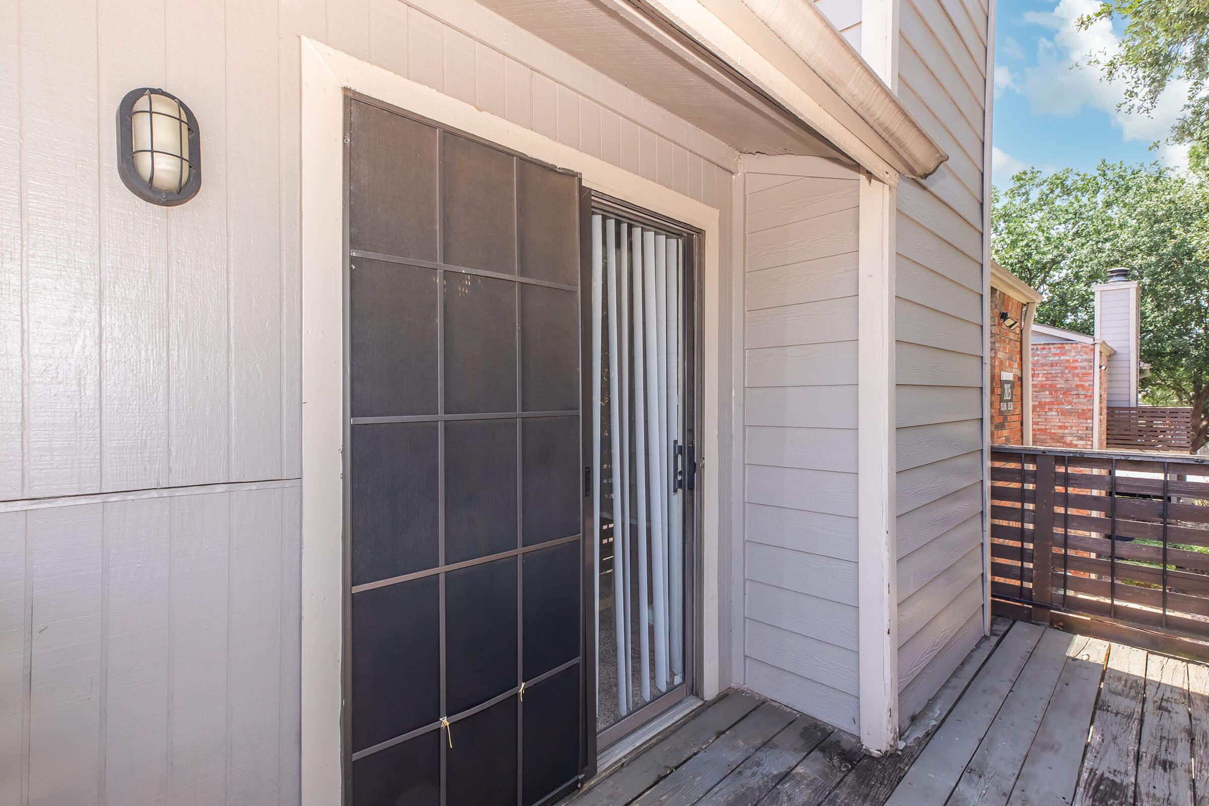 A sliding glass door with vertical blinds leads onto a wooden deck, set against a light gray exterior wall. A wall-mounted light fixture is beside the door, and greenery is visible beyond the deck. The scene is well-lit, suggesting a sunny day.