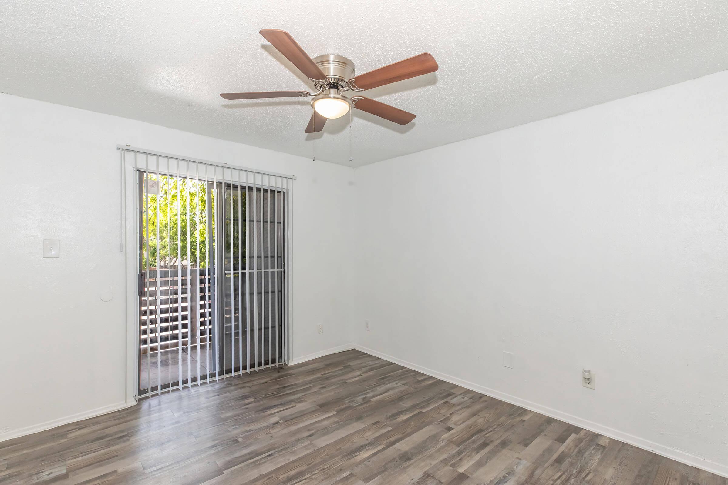 A light and airy empty room featuring a ceiling fan, light grey walls, and laminate flooring. There are sliding glass doors with vertical blinds leading to an outdoor area, allowing natural light to enter the space. The overall ambiance is clean and simple, suitable for personalization or decoration.