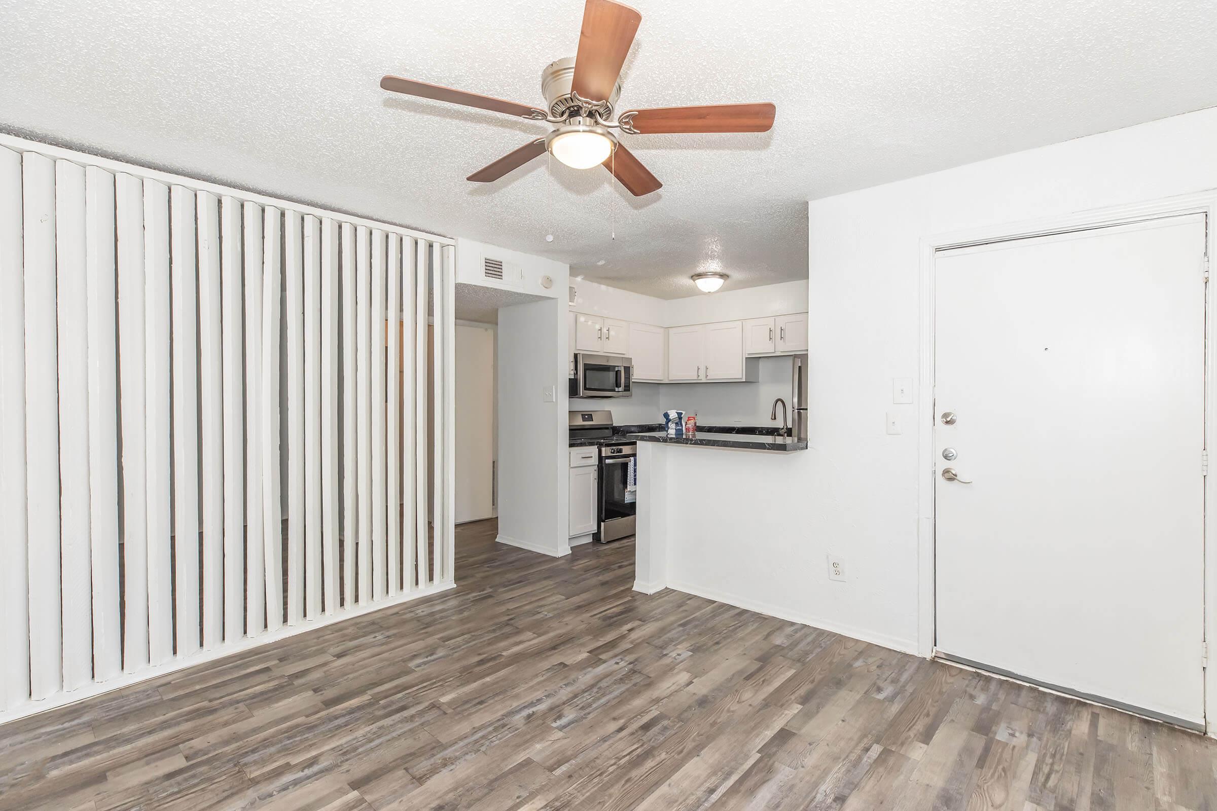 A modern open-concept living space featuring a wooden floor, a ceiling fan, and white walls. The kitchen is visible with stainless steel appliances and a countertop. A door leads outside, and there are vertical blinds on one wall, contributing to a light and airy atmosphere.