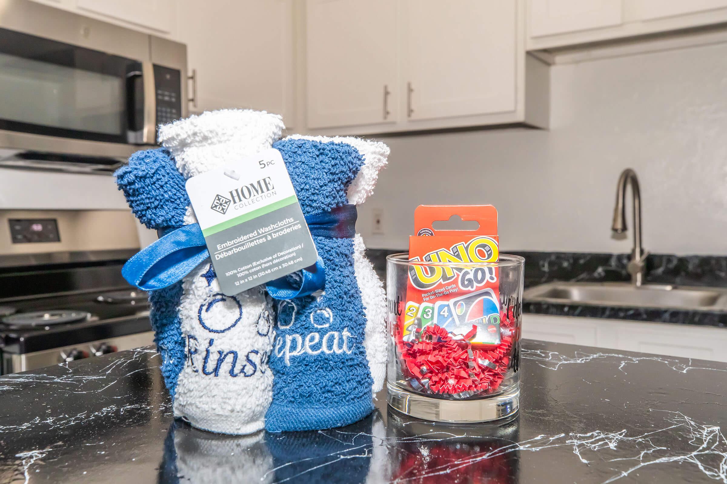A small kitchen countertop displaying a blue and white towel set labeled "Home Essentials" with a tag, beside a tumbler glass filled with red crinkle paper and a pack of Uno Go cards. The background features a modern kitchen with a black countertop, microwave, and sink.