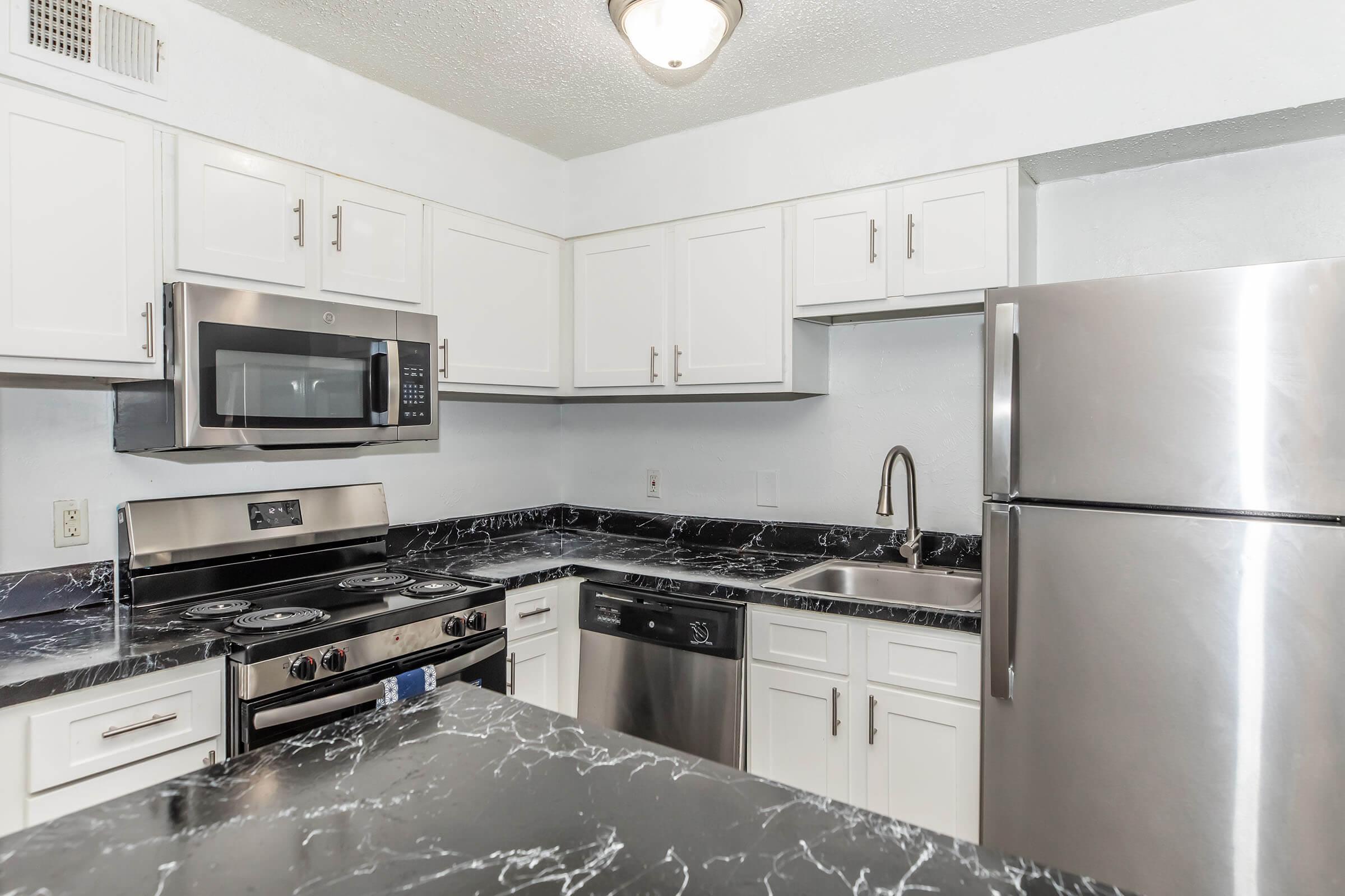 Modern kitchen featuring white cabinets, a stainless steel refrigerator, a sleek black marble countertop, and a combination microwave and oven. Includes a stainless steel sink and a dishwasher, with an open layout that enhances the space's functionality and style.