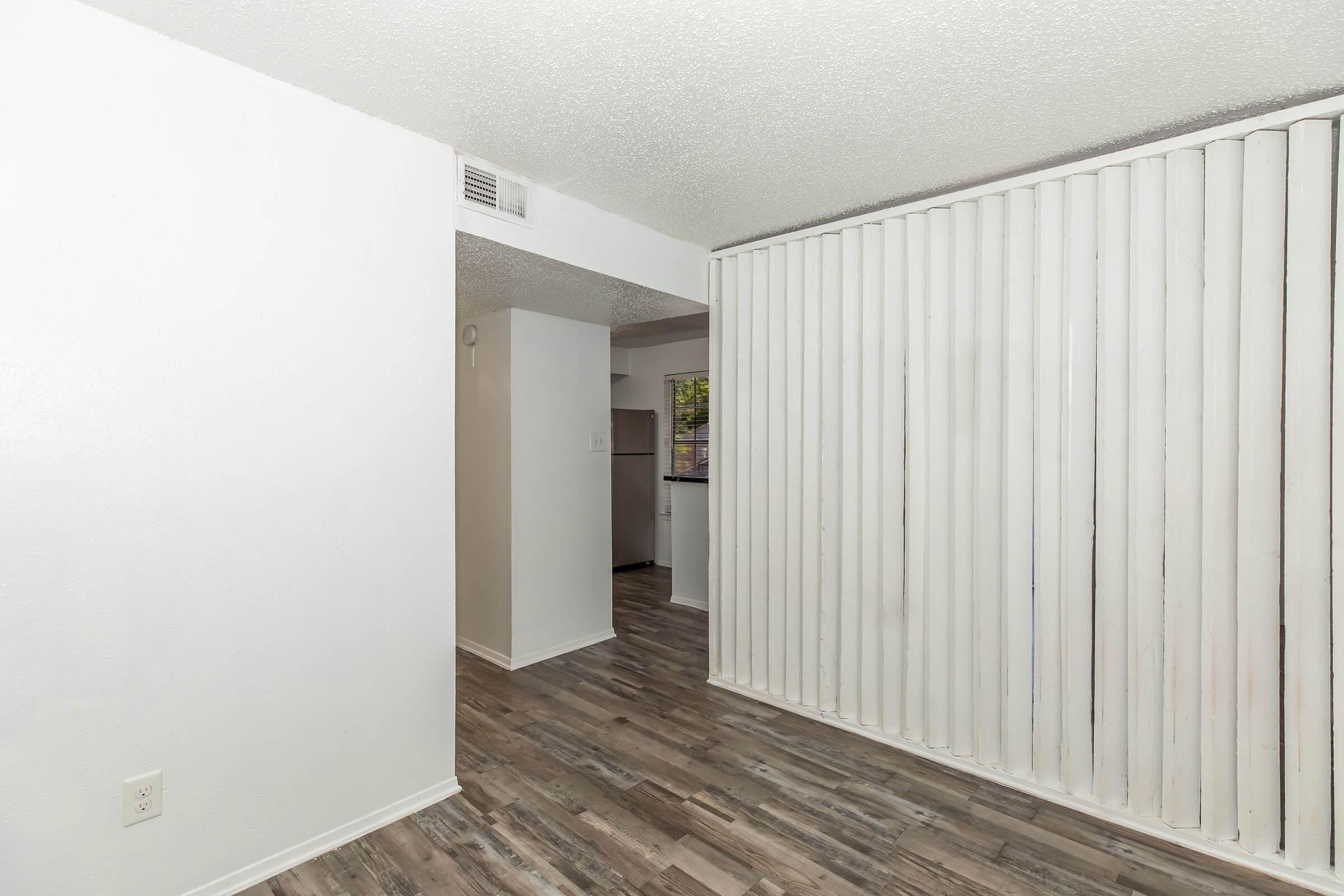 Interior view of a room with light-colored walls and wood-look flooring. A white panelled divider can be seen, separating the space. In the background, a kitchen area is partially visible. Natural light enters through the windows, creating a bright atmosphere.