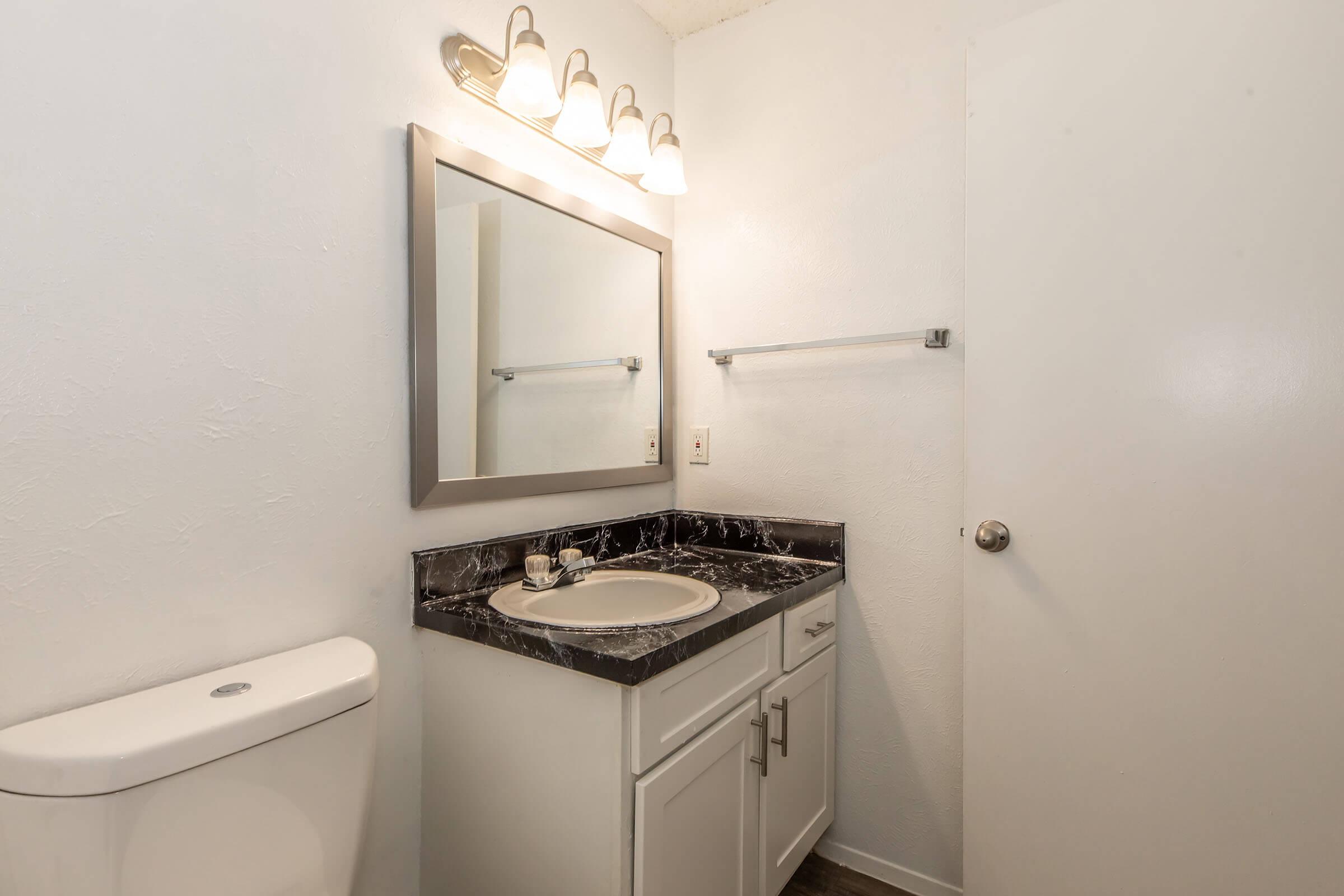A small bathroom featuring a white toilet, a sink with a black and white marble countertop, and a framed mirror above it. The wall is painted white, and there is a towel rack. A light fixture with multiple bulbs is mounted above the mirror, providing illumination. The door is partially visible on the right.