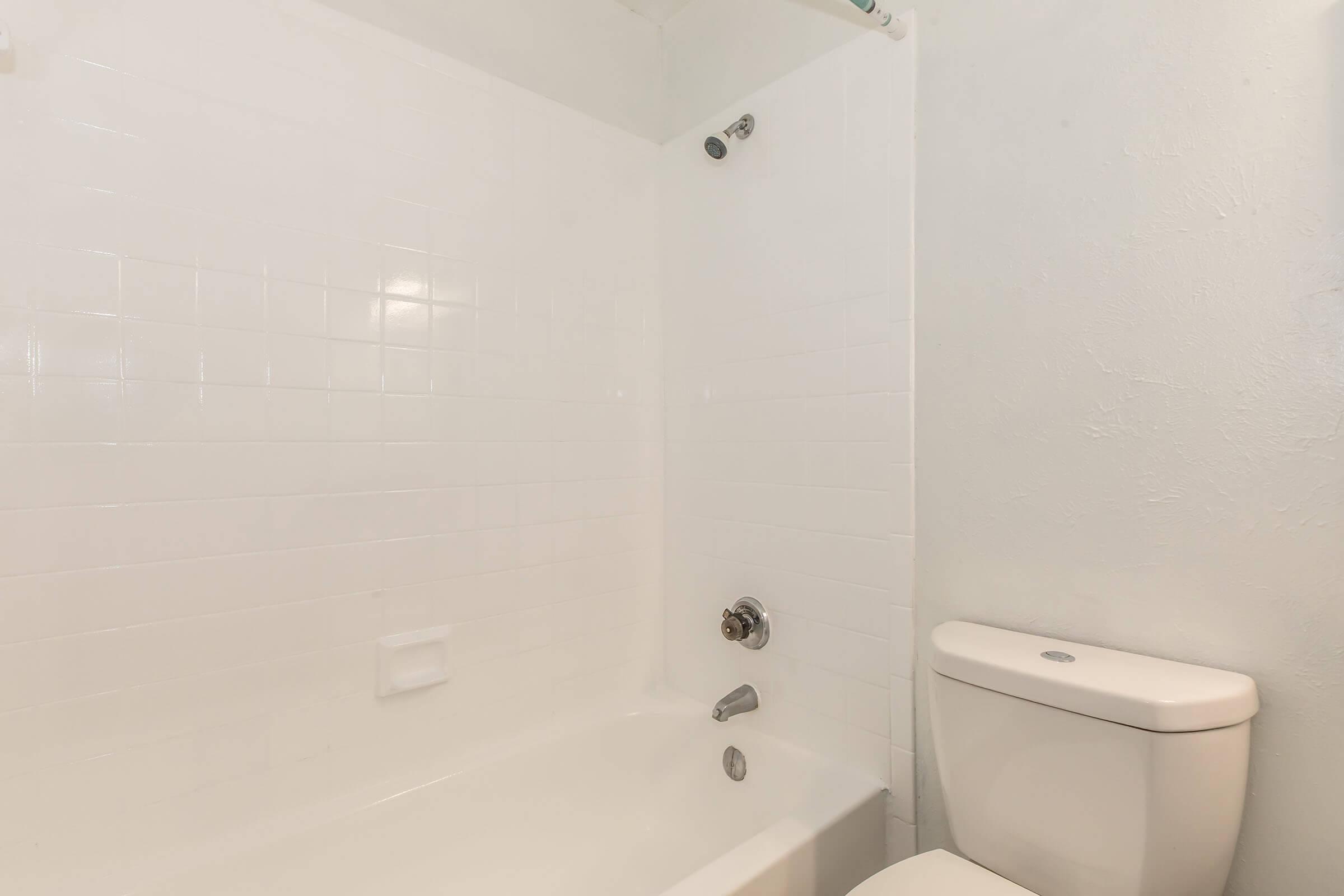 A clean, minimalistic bathroom featuring a white tiled shower and bathtub combination with a chrome showerhead, a wall-mounted faucet, and a white toilet nearby. The walls are painted in a light color, enhancing the bright and airy feel of the space.