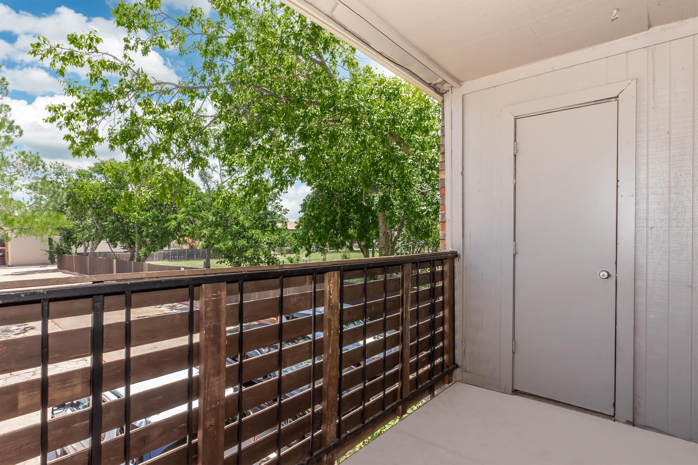 A balcony view featuring wooden railing and a closed door. Lush green trees are visible in the background, with a clear blue sky and fluffy clouds overhead. The setting offers a calm and inviting atmosphere.