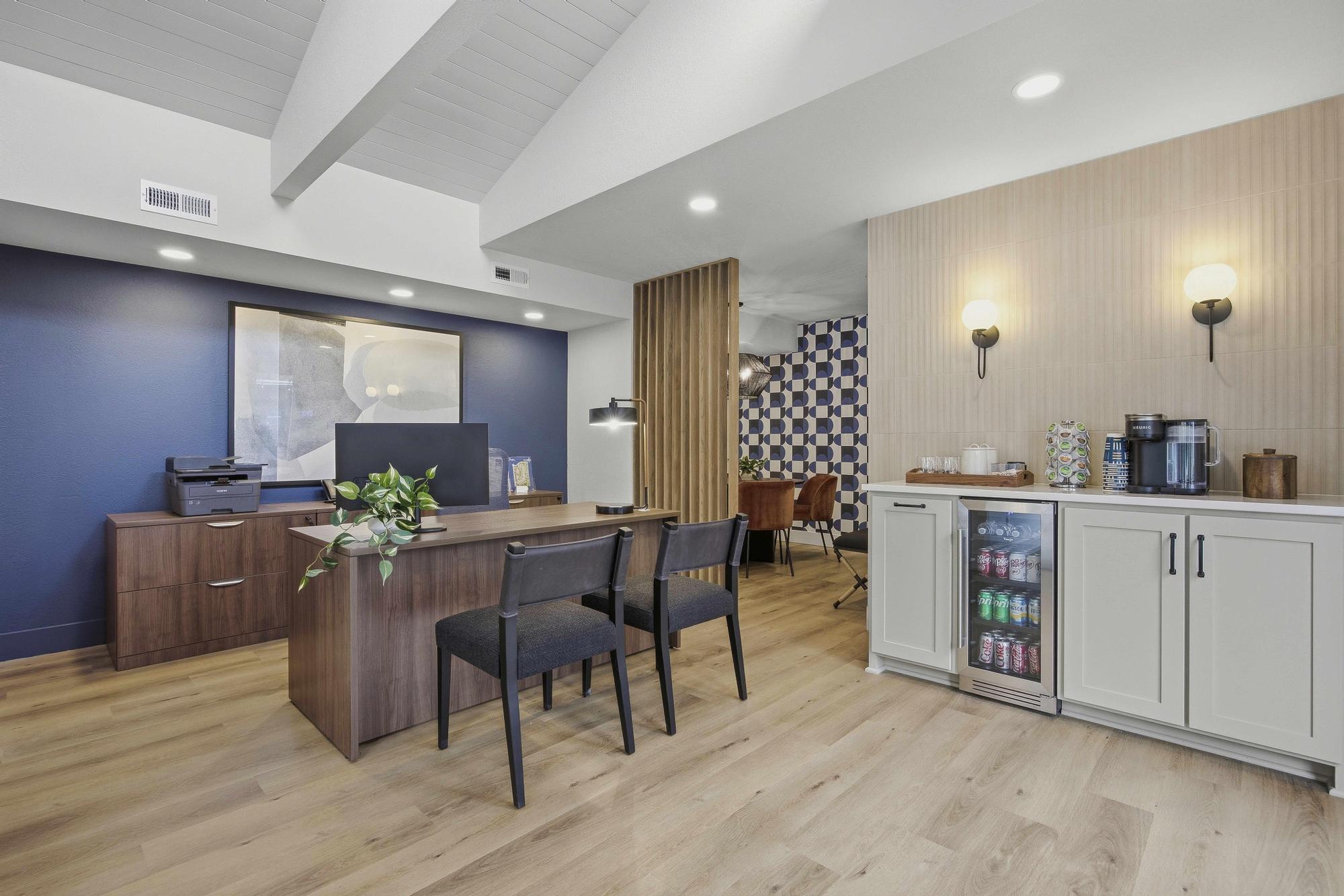 A modern office space with a wooden desk and two black chairs. A printer is positioned on the desk, and a small kitchenette area features a glass-front refrigerator stocked with drinks. The walls are painted in contrasting colors, with decorative elements and a large framed artwork in the background.