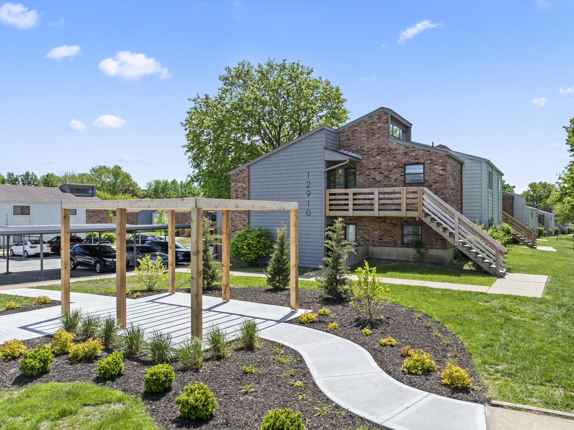 A view of a landscaped residential area featuring a multi-unit brick and gray building. In the foreground, there is a wooden pergola surrounded by neatly arranged flower beds and green grass. The sky is bright blue with a few clouds, and parking spaces are visible in the background.