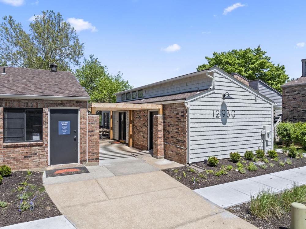 Exterior view of a modern building with a brick facade and a light-colored siding extension. A pathway leads to a covered entrance, flanked by greenery. The sky is clear and blue, with a few trees in the background. The building has a contemporary design and a welcoming atmosphere.