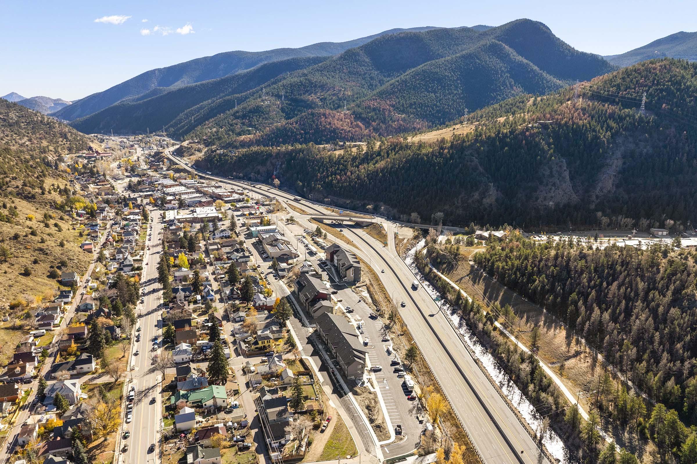 Aerial view of a small town nestled between mountains, featuring a highway running through the landscape. Below, various buildings and homes are visible, along with patches of trees displaying autumn colors. The expansive mountainous backdrop enhances the scenic beauty of the area.