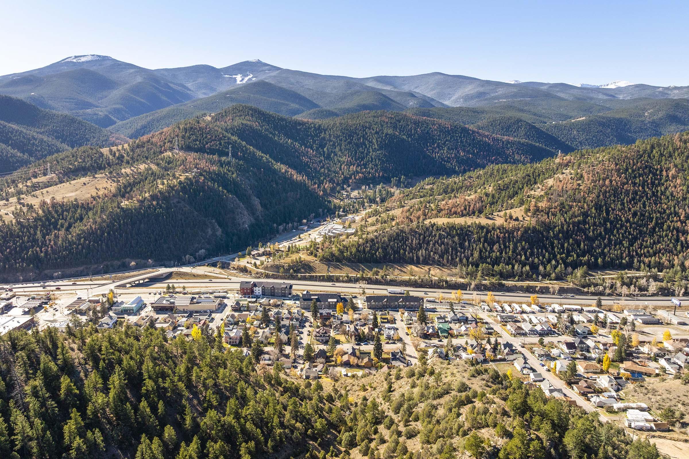 Aerial view of a small mountain town nestled in a valley surrounded by rolling hills and peaks. The town features a mix of buildings and businesses, with a highway running through it. Autumn foliage adds splashes of color to the landscape, highlighting the natural beauty of the area.